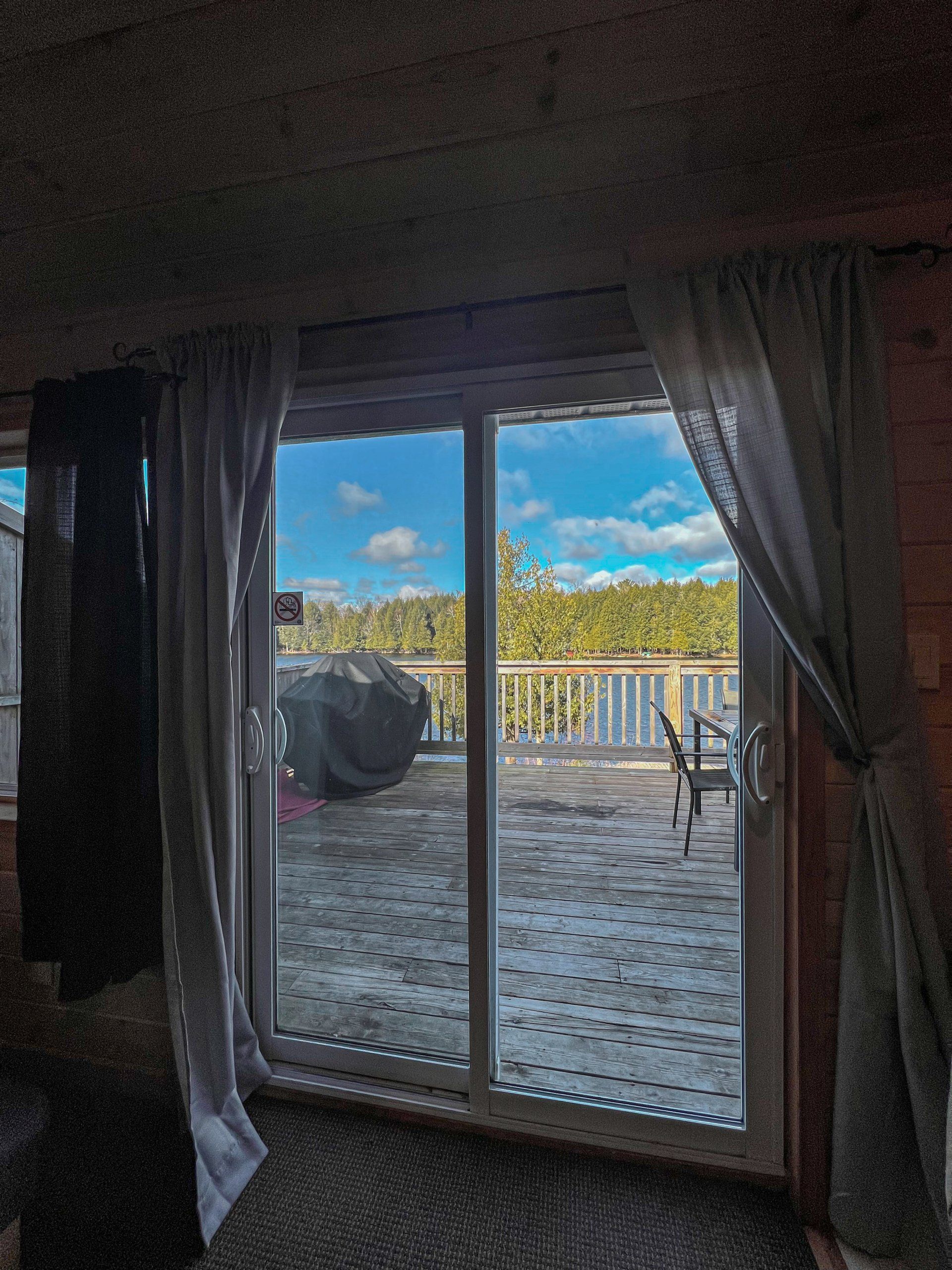 View through sliding glass doors to a sunny deck with white railing, blue sky, and trees beyond.