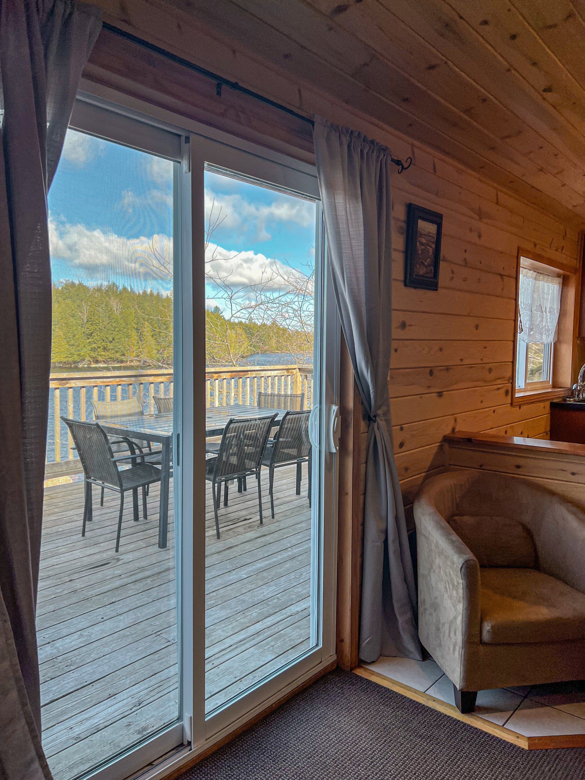 Cabin interior with sliding glass door opening to a deck with chairs and a lake view