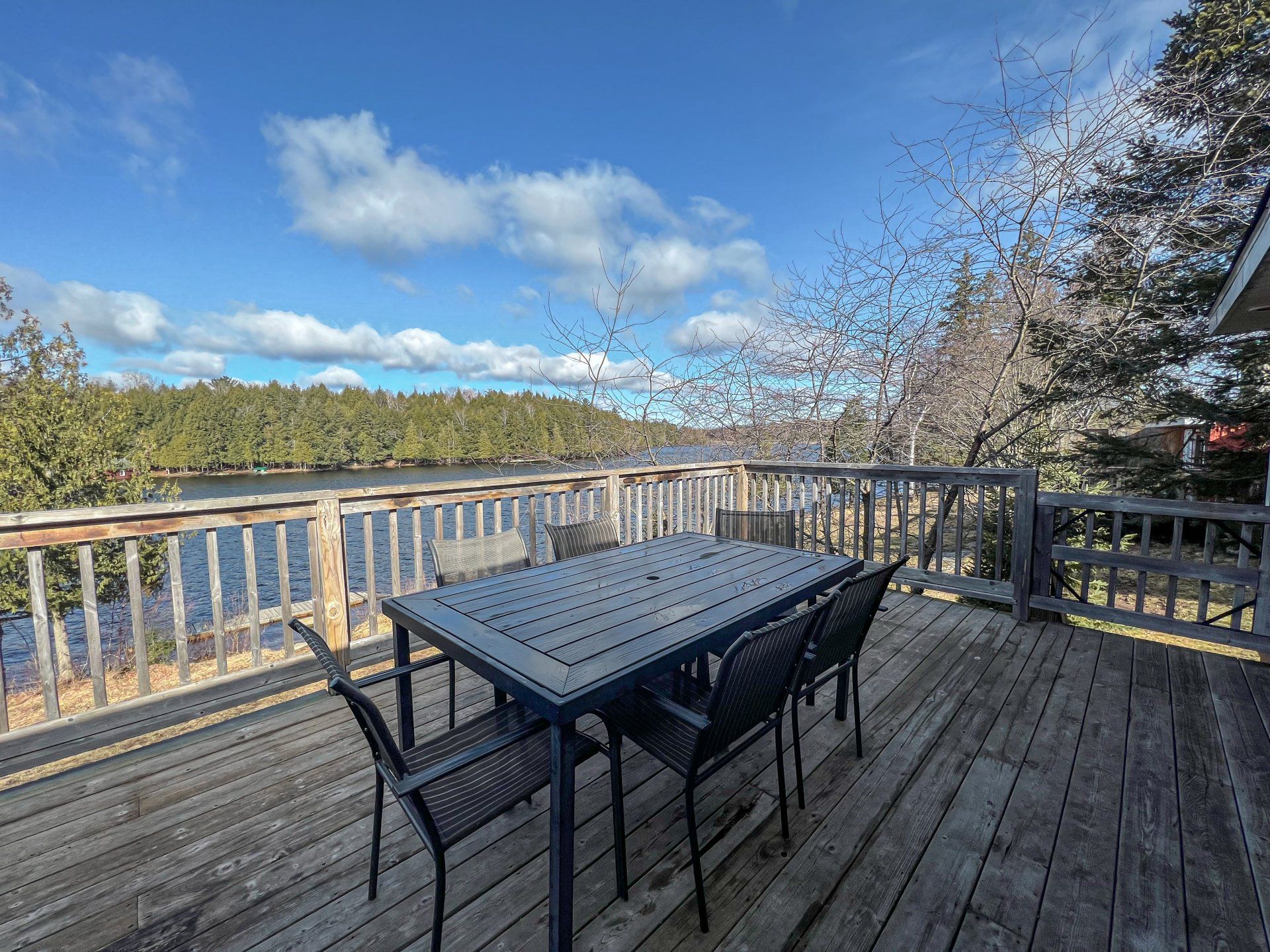 Wooden deck with black patio table and chairs overlooking a lake and trees under a blue sky