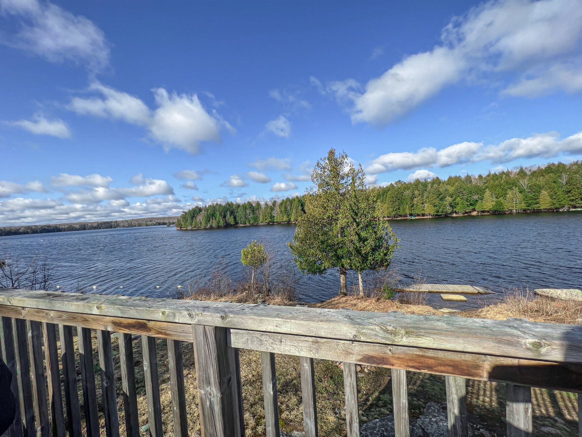 Wooden fence overlooking a blue lake with small trees, green shoreline, and a cloudy sky