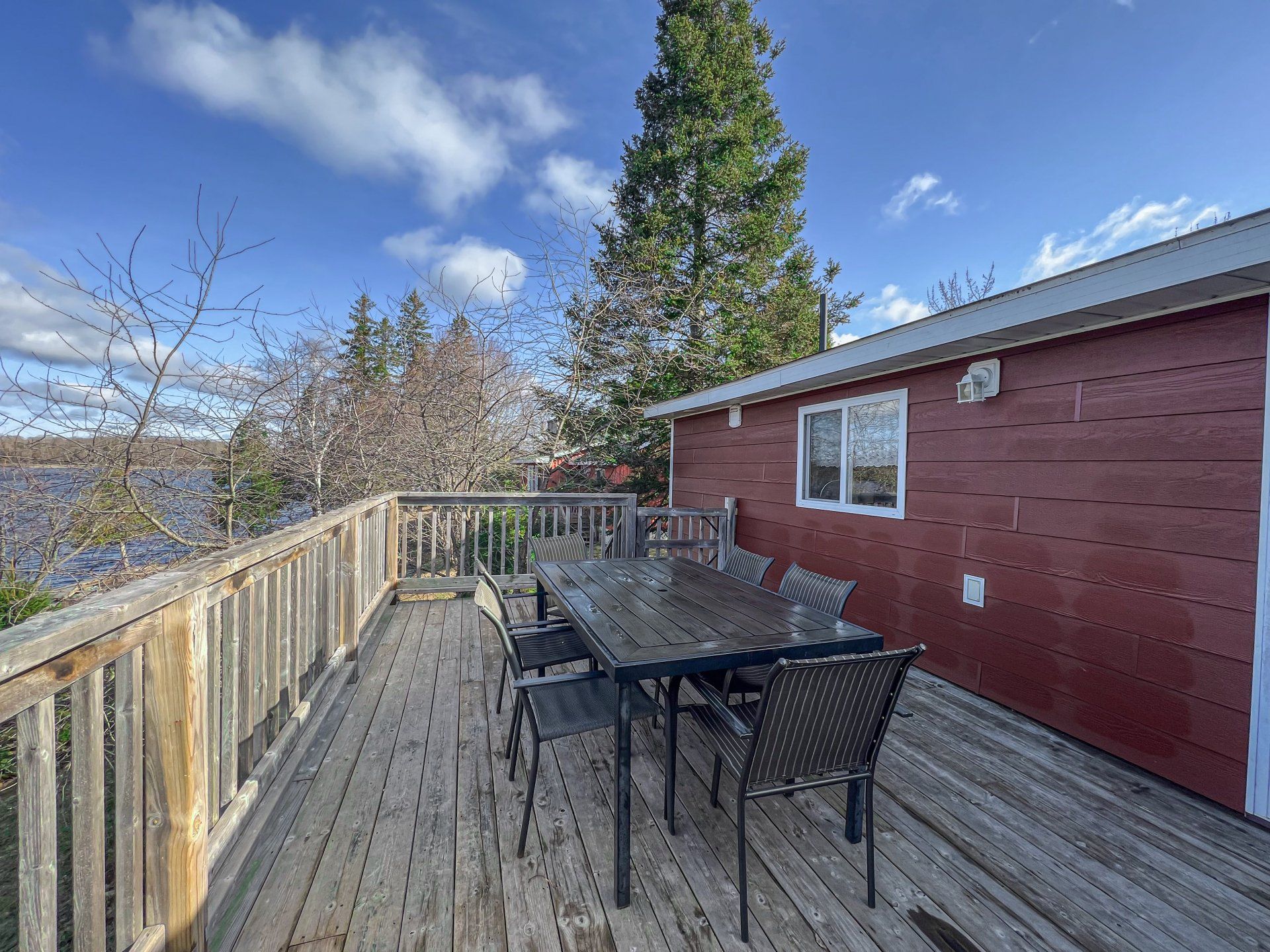 Wooden deck with patio table and chairs beside a red house, overlooking trees and a lake under a blue sky