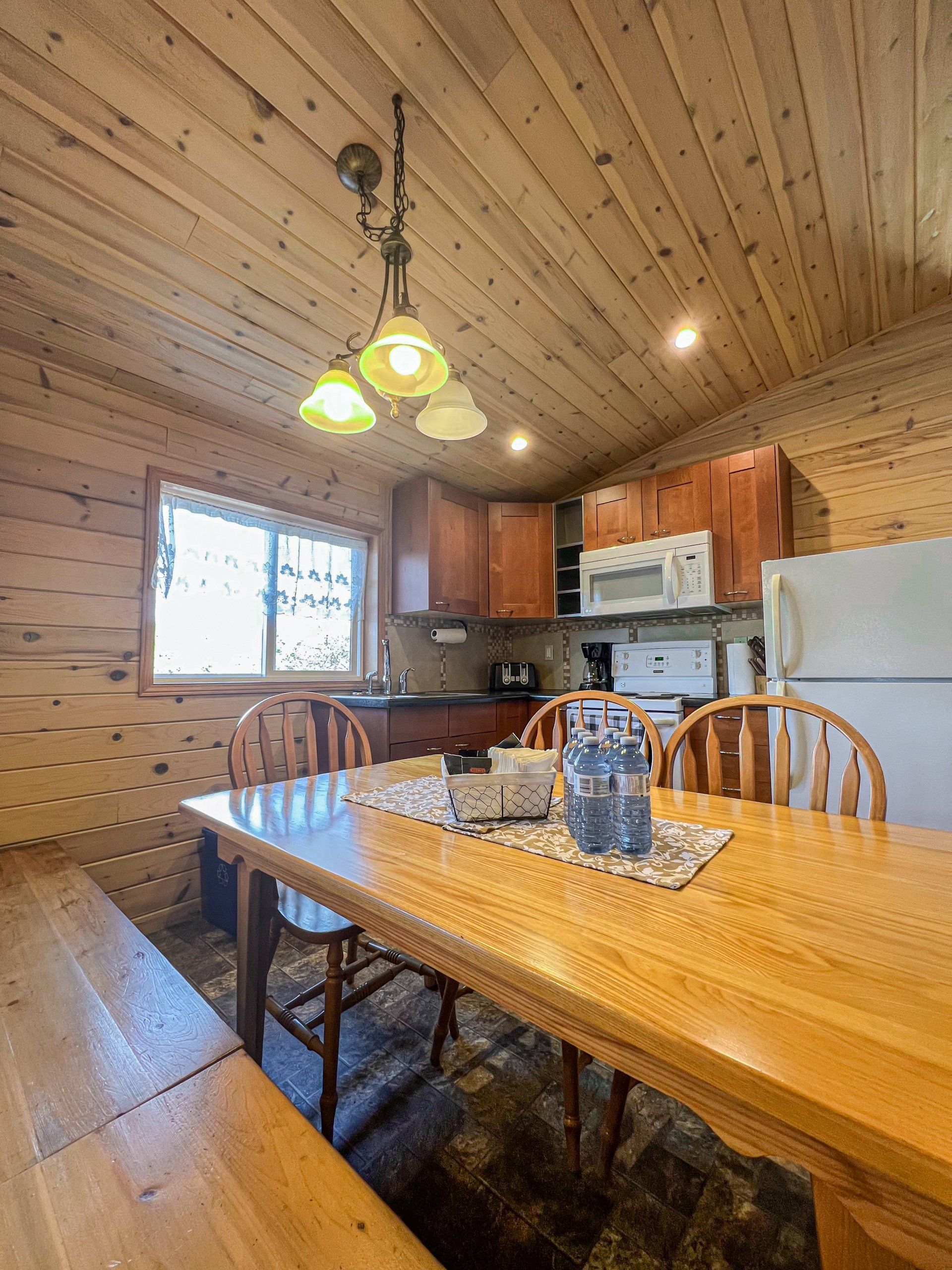 Wood-paneled dining room with a long wooden table, hanging lights, and a kitchen in the background.