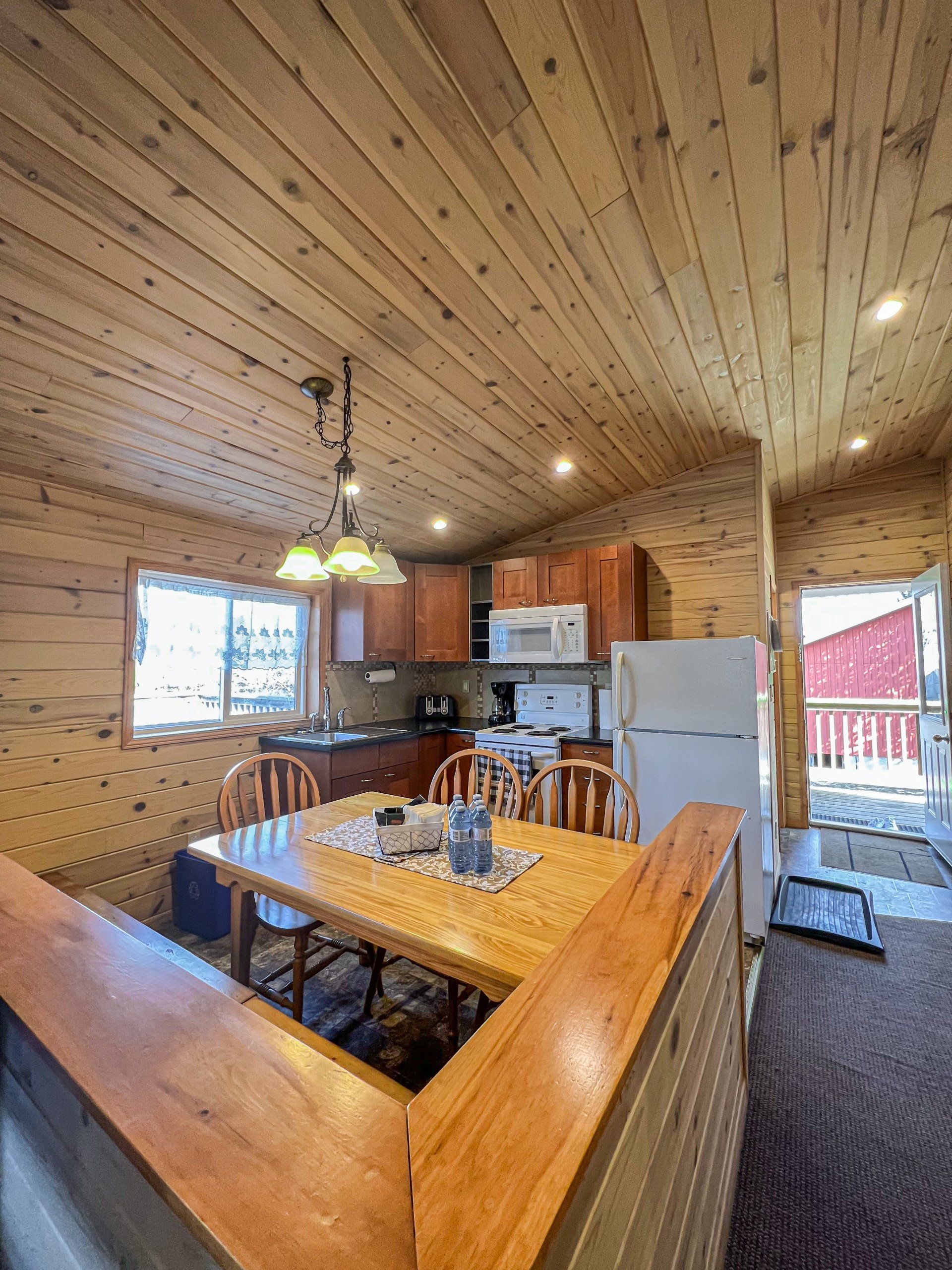 Wood-paneled cabin kitchen and dining area with wooden table, chairs, white appliances, and large windows