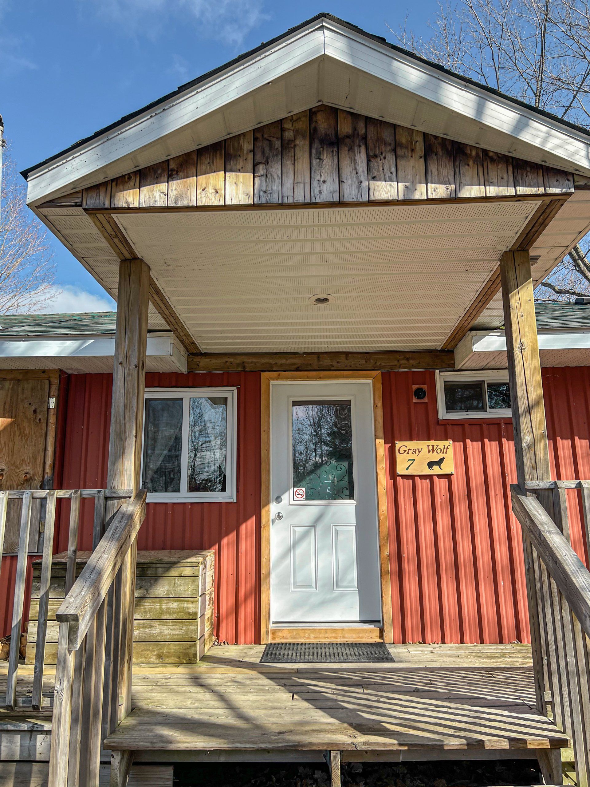Small red cabin porch with white door, wooden steps, and a peaked roof in bright sunlight