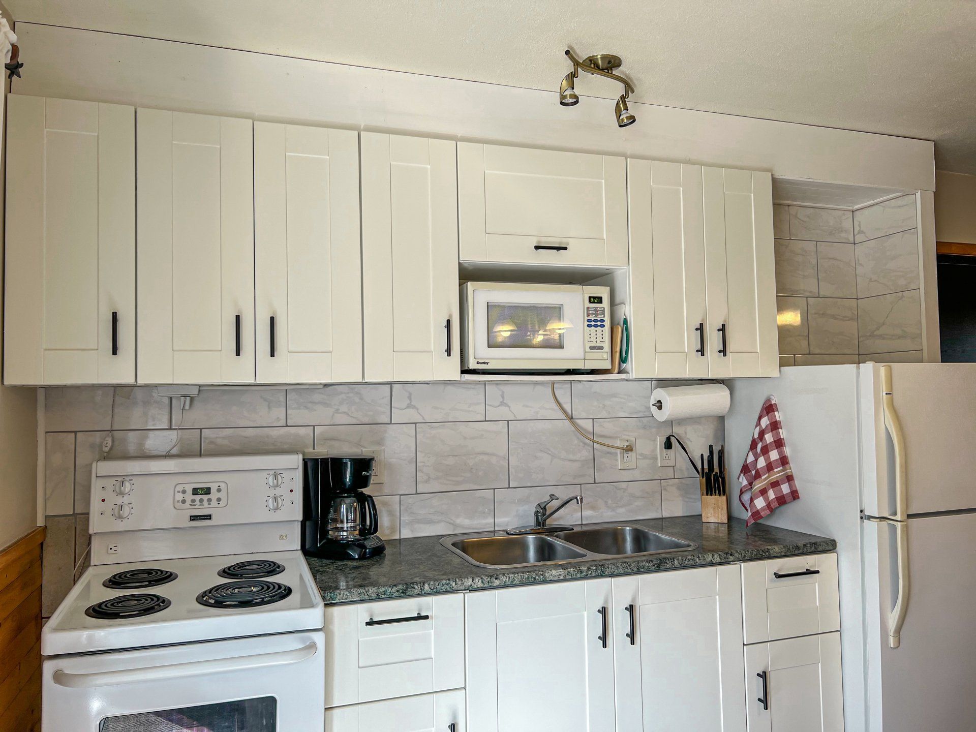 Small white kitchen with cabinets, stove, sink, microwave, and refrigerator, lit by sunlight.