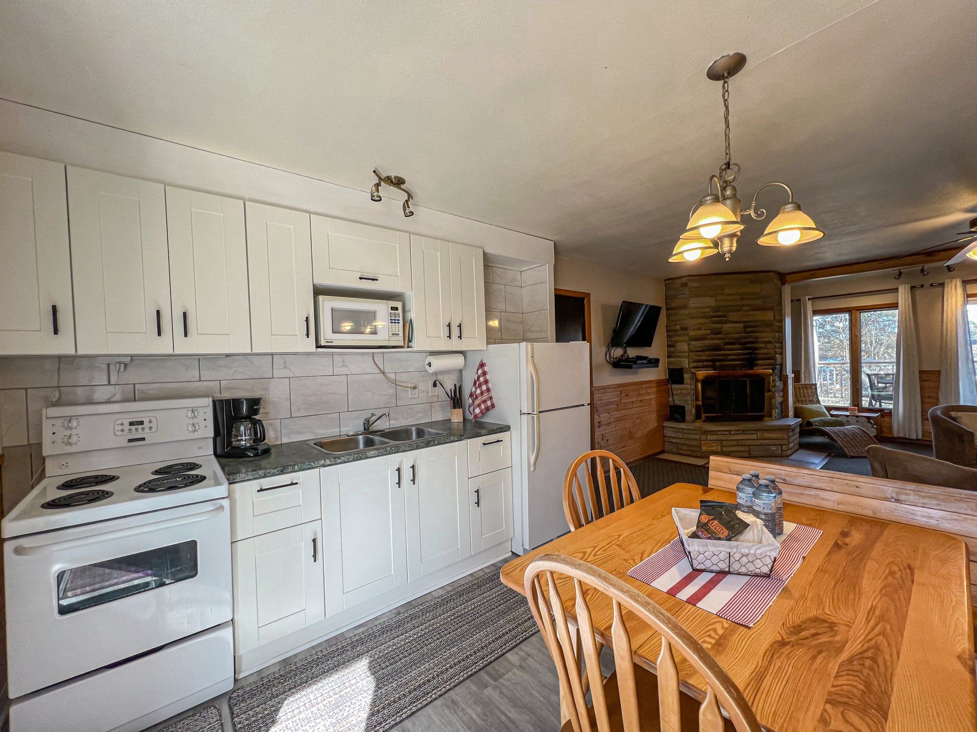 Bright kitchen and dining area with white cabinets, stove, wooden table, and stone fireplace in the background