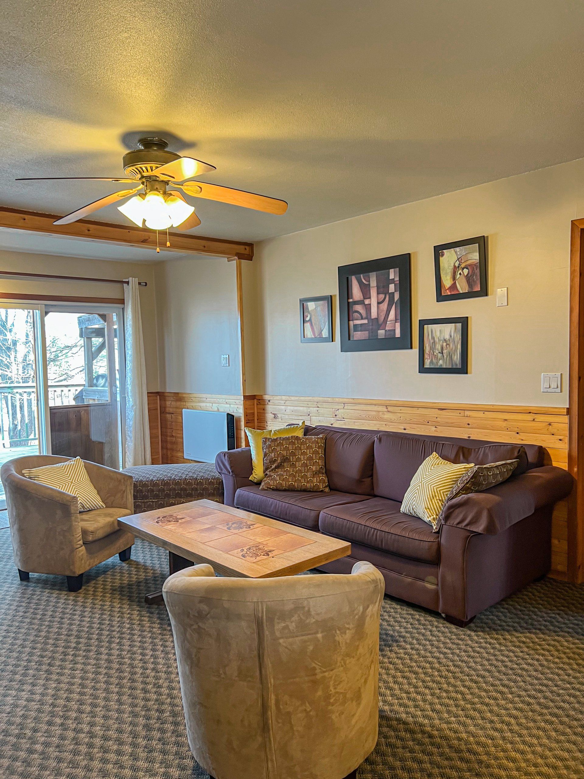 Cozy living room with brown sofa, tan chairs, ceiling fan, and framed wall art by a window.