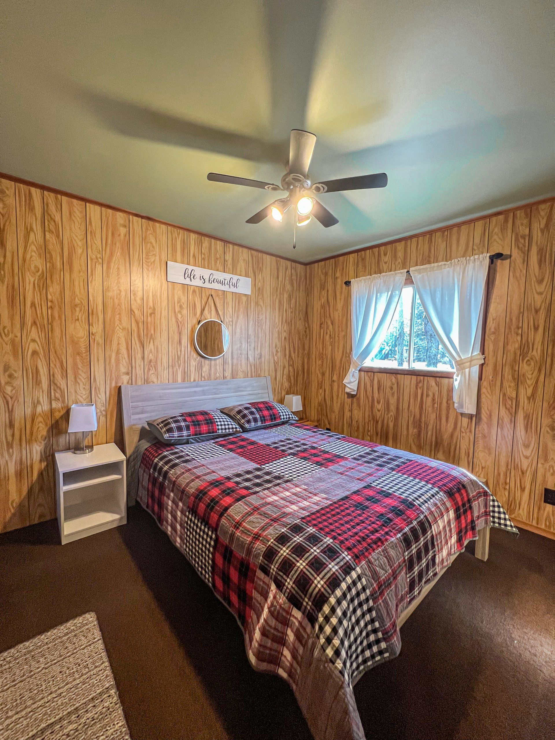 Bedroom with a plaid bedspread, wood-paneled walls, ceiling fan, and a window with curtains.