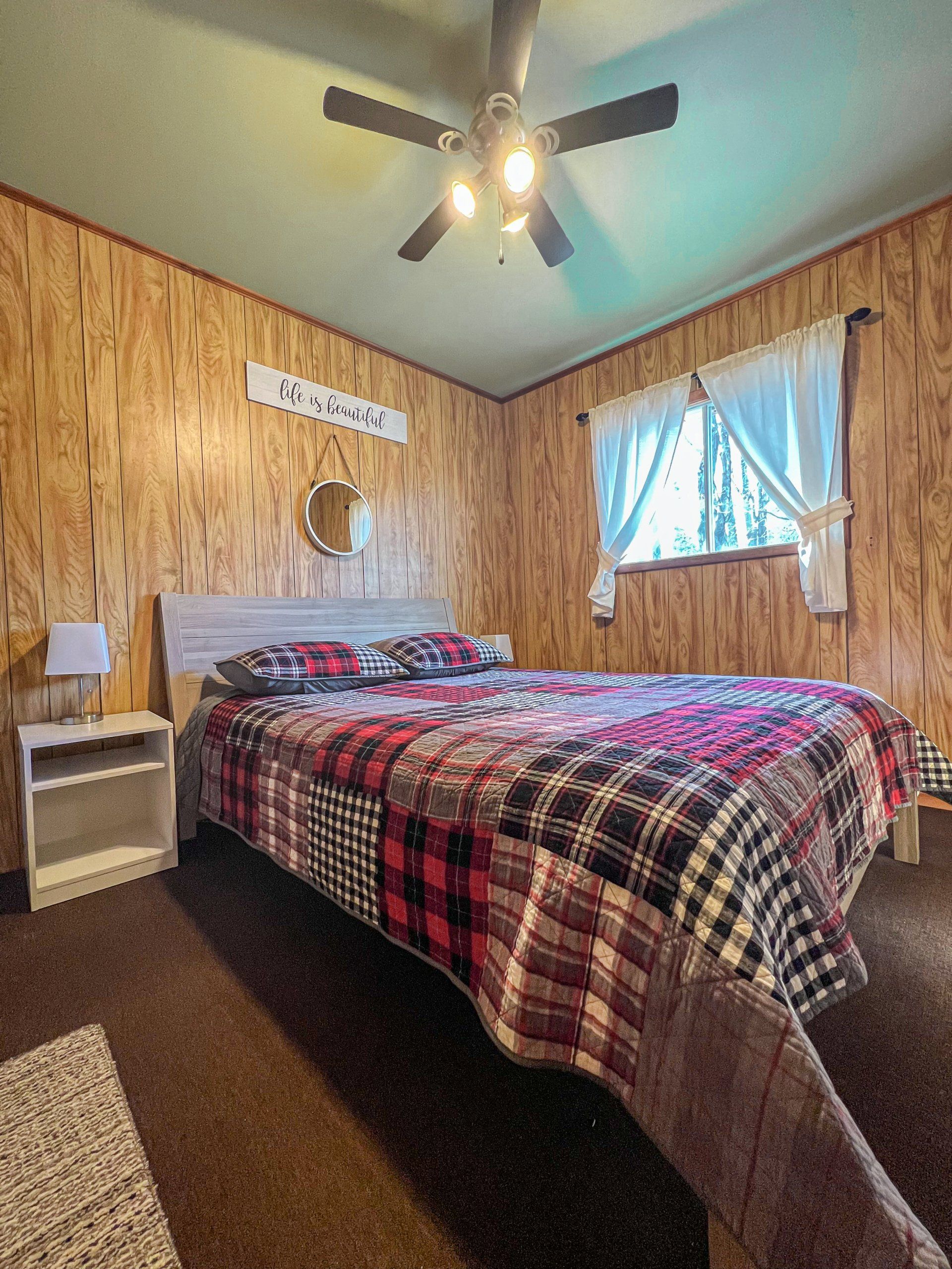 Small bedroom with wood-paneled walls, plaid bedspread, ceiling fan, and sunlit window with blue curtains