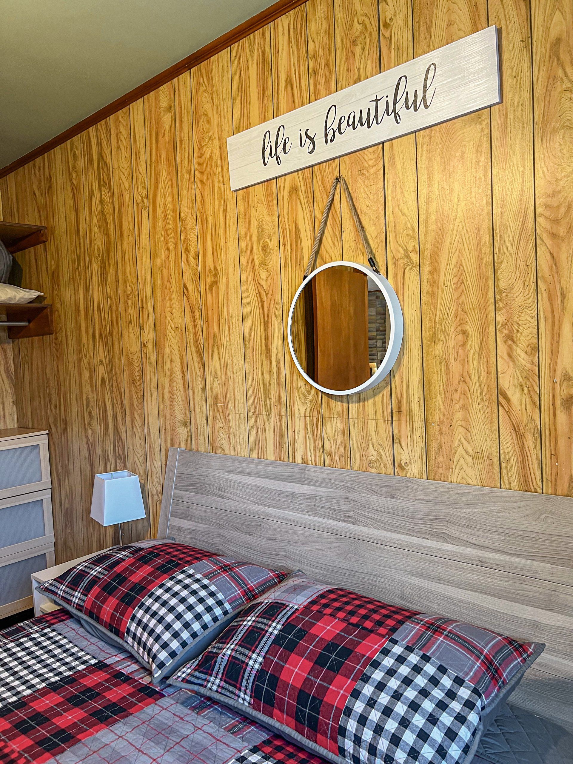 Cozy bed with red plaid bedding beneath a round mirror and rustic wood wall sign reading “His & Her’s.”
