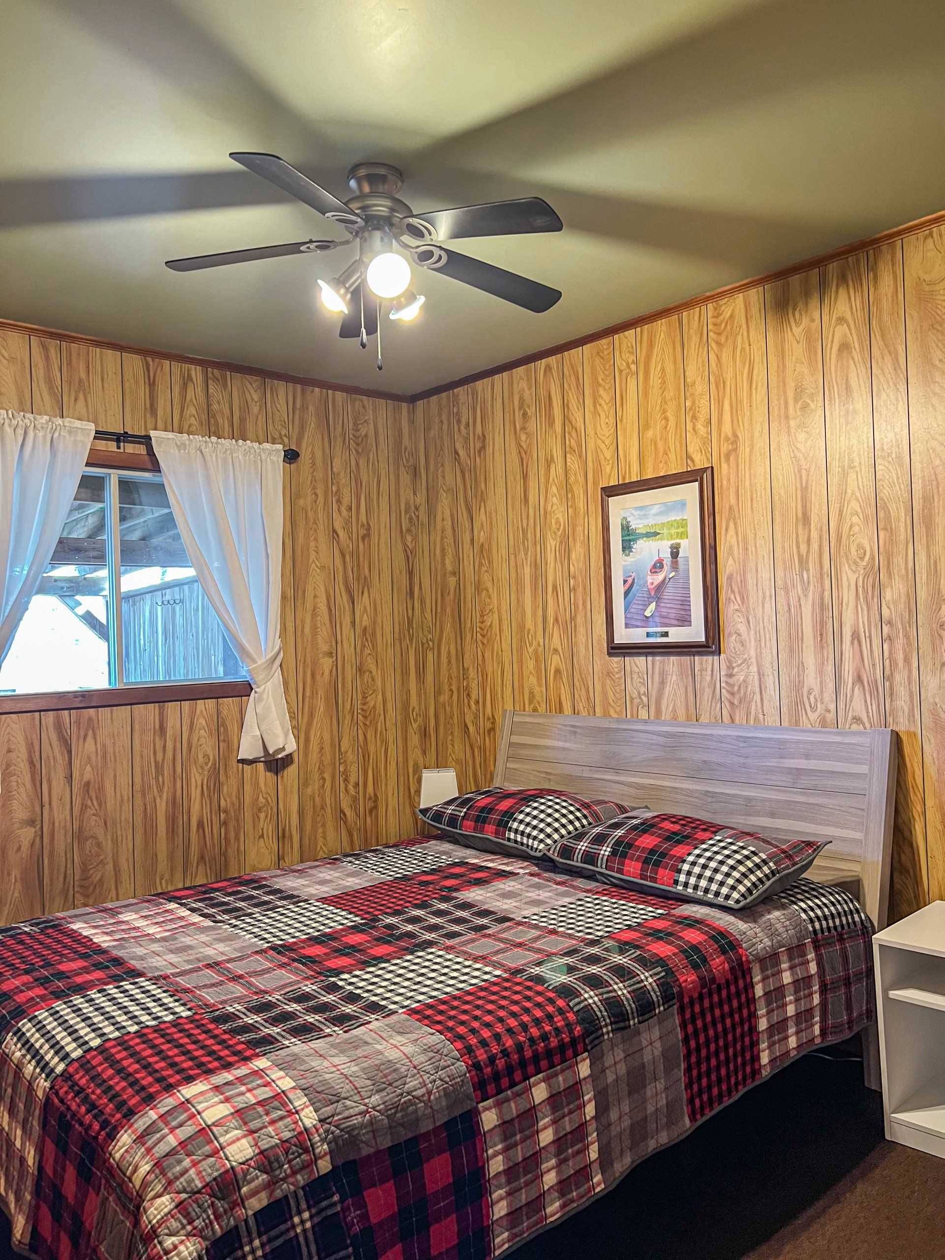 Cozy bedroom with wood-paneled walls, plaid bedspread, ceiling fan, and a window with curtains.