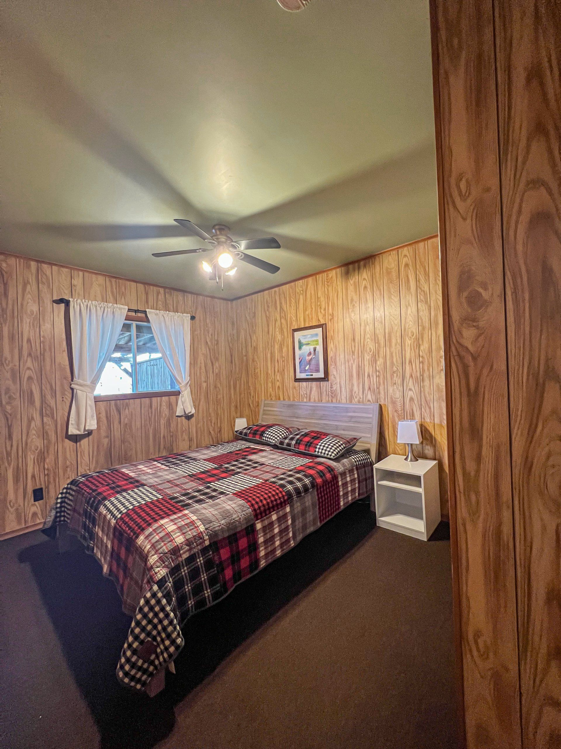 Cozy bedroom with wood-paneled walls, plaid bedspread, ceiling fan, and small window.