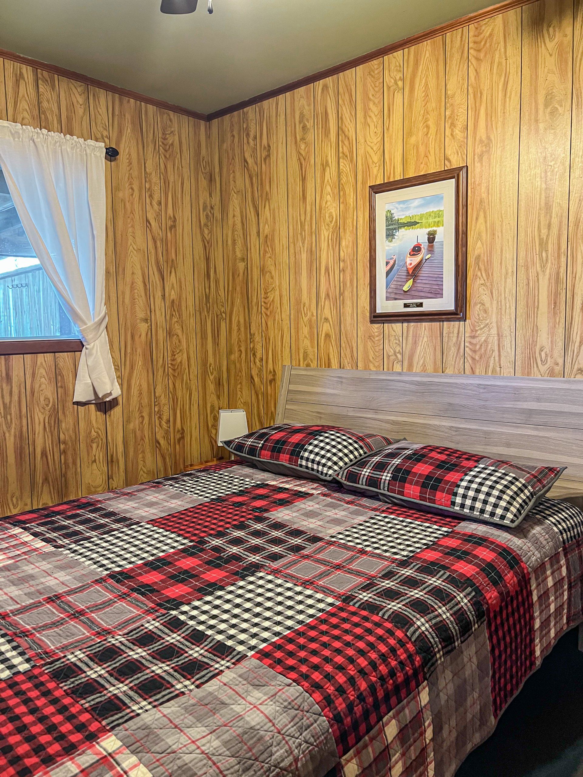 Cozy bedroom with plaid bedspread, wood-paneled walls, window curtains, and framed wall art.