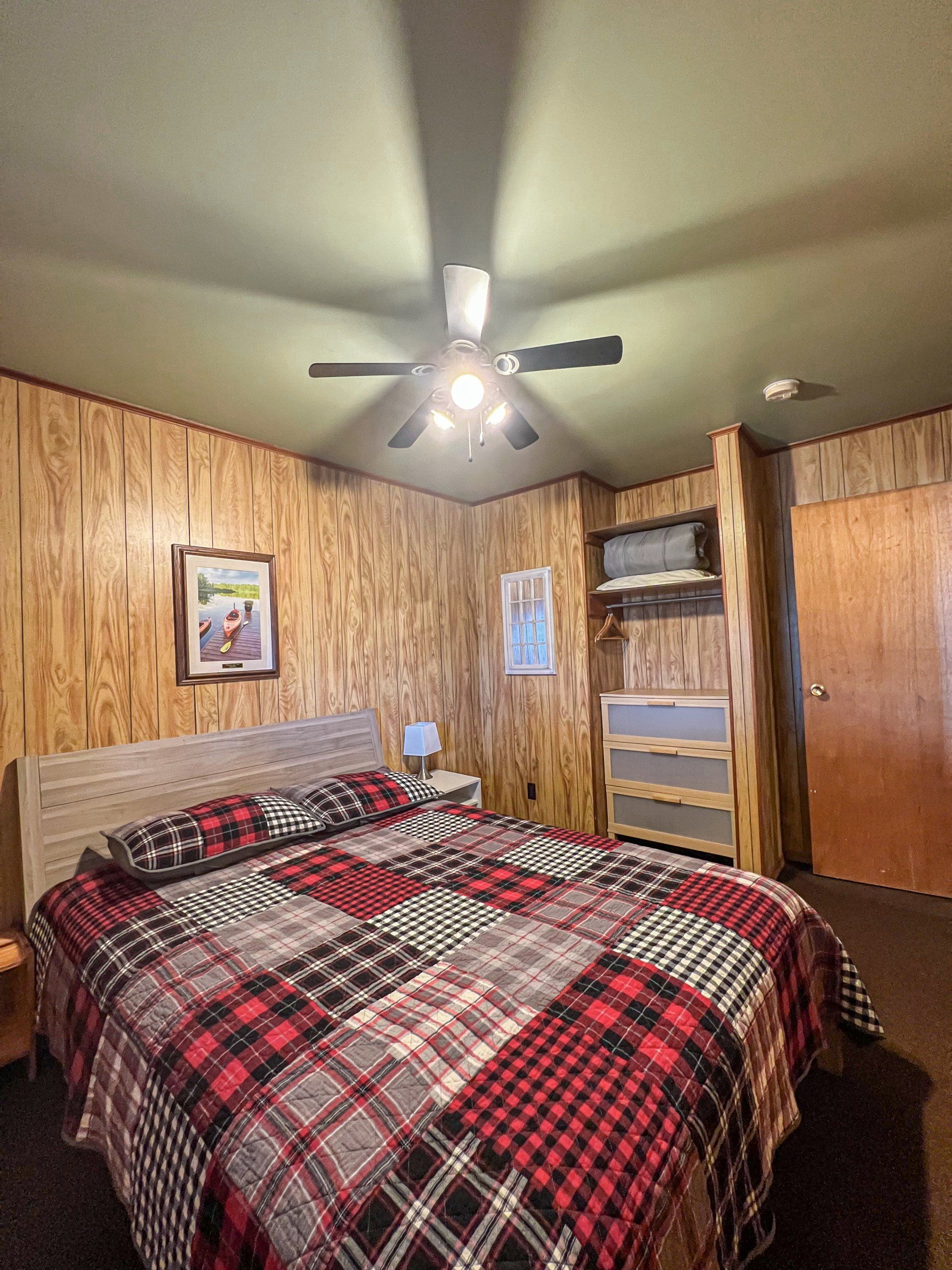 Cozy bedroom with plaid bedspread, wood-paneled walls, ceiling fan, and dresser with mirror