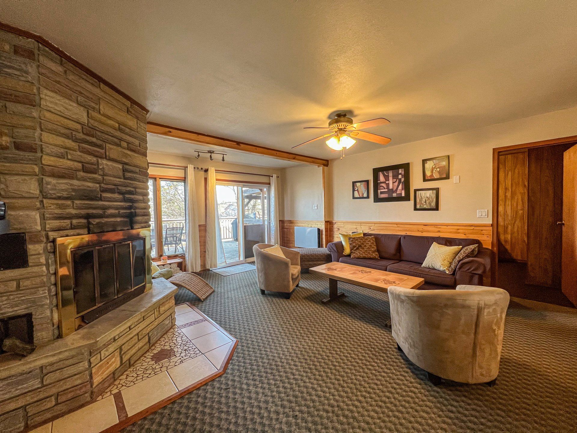 Cozy living room with stone fireplace, sectional sofa, armchairs, patterned carpet, and ceiling fan.