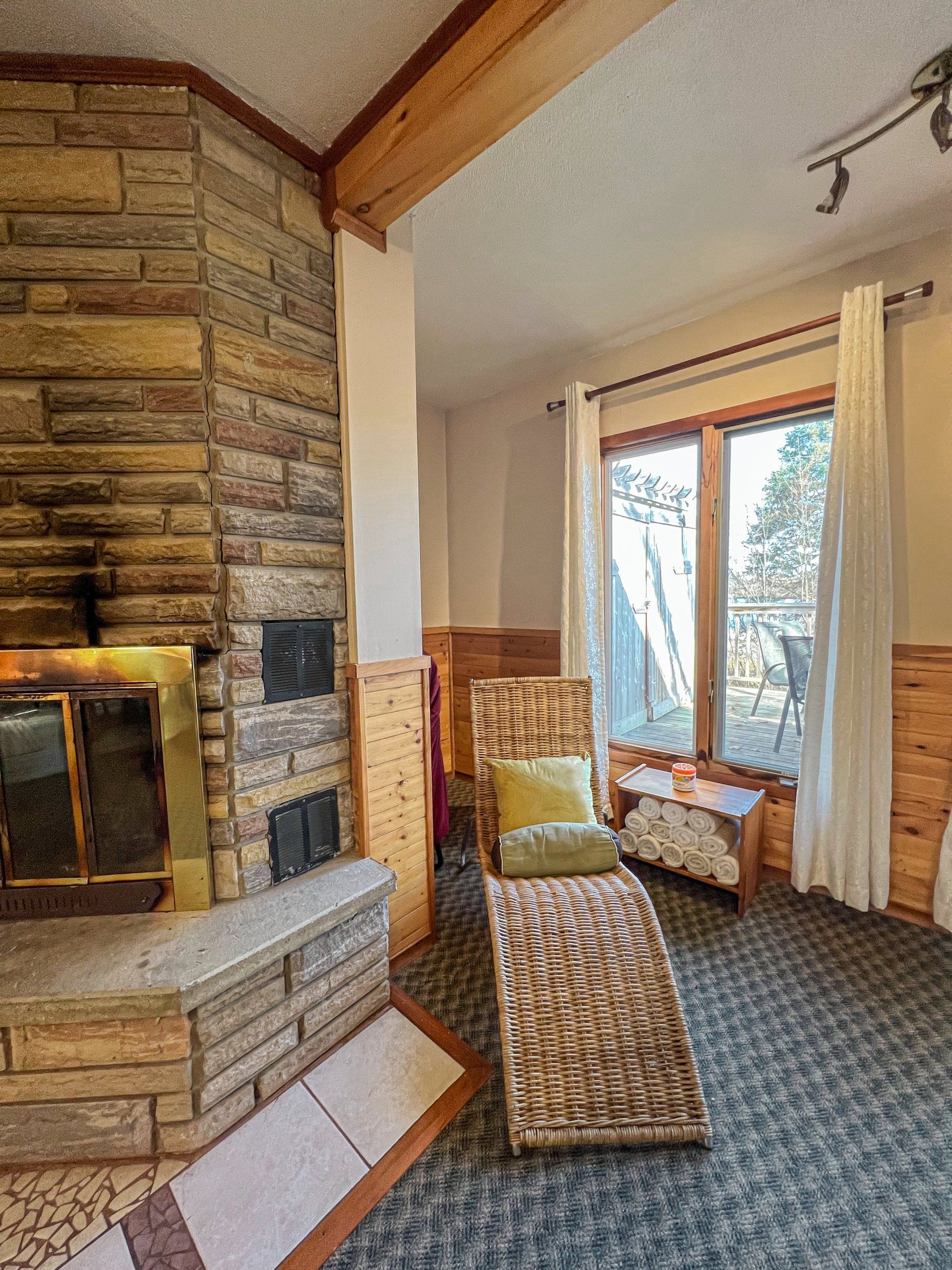 Stone fireplace in a cozy living room with wicker chairs and a large window overlooking trees.