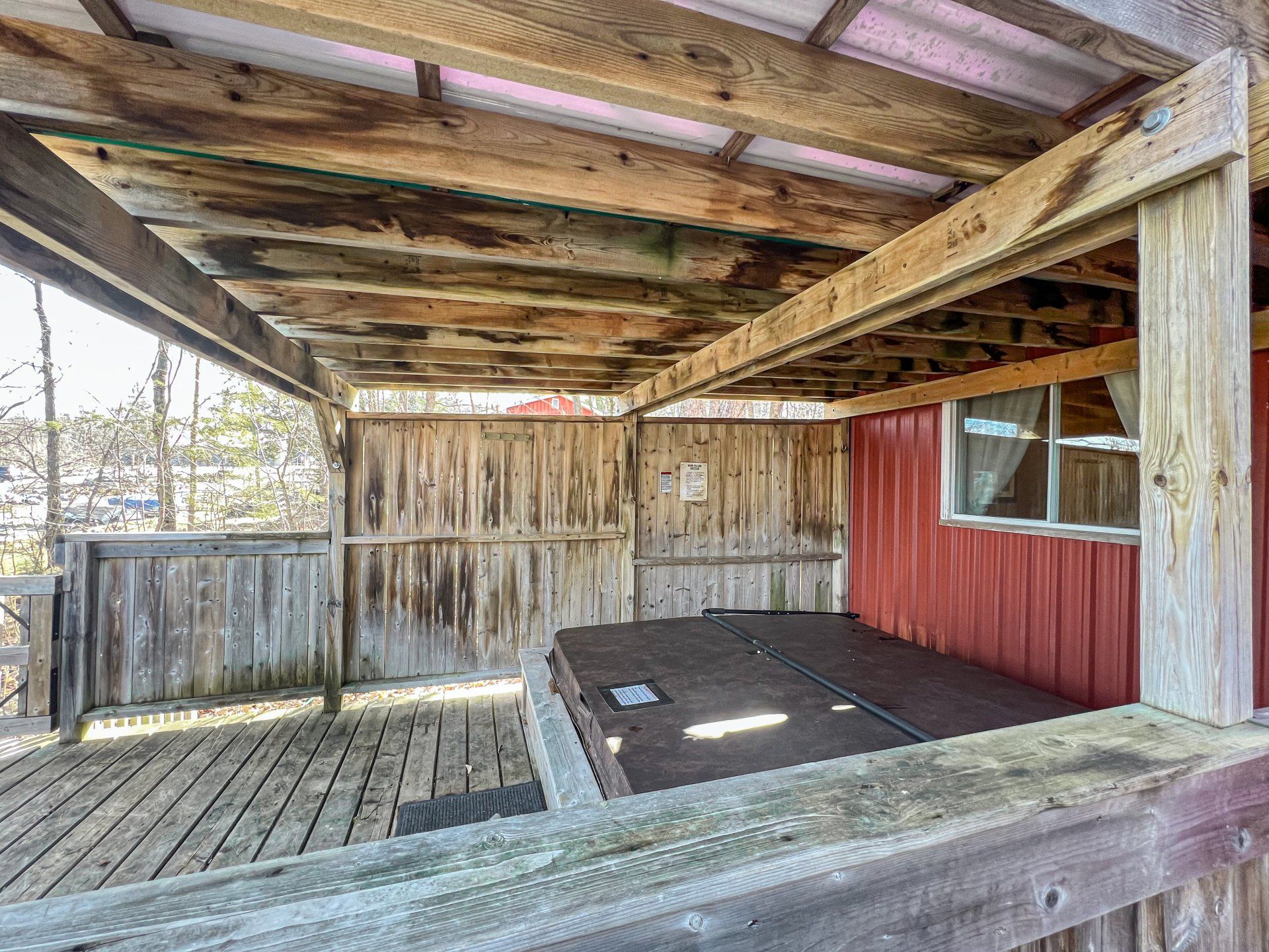 Covered wooden deck with weathered railing, red siding, and a hot tub underneath a slatted roof.