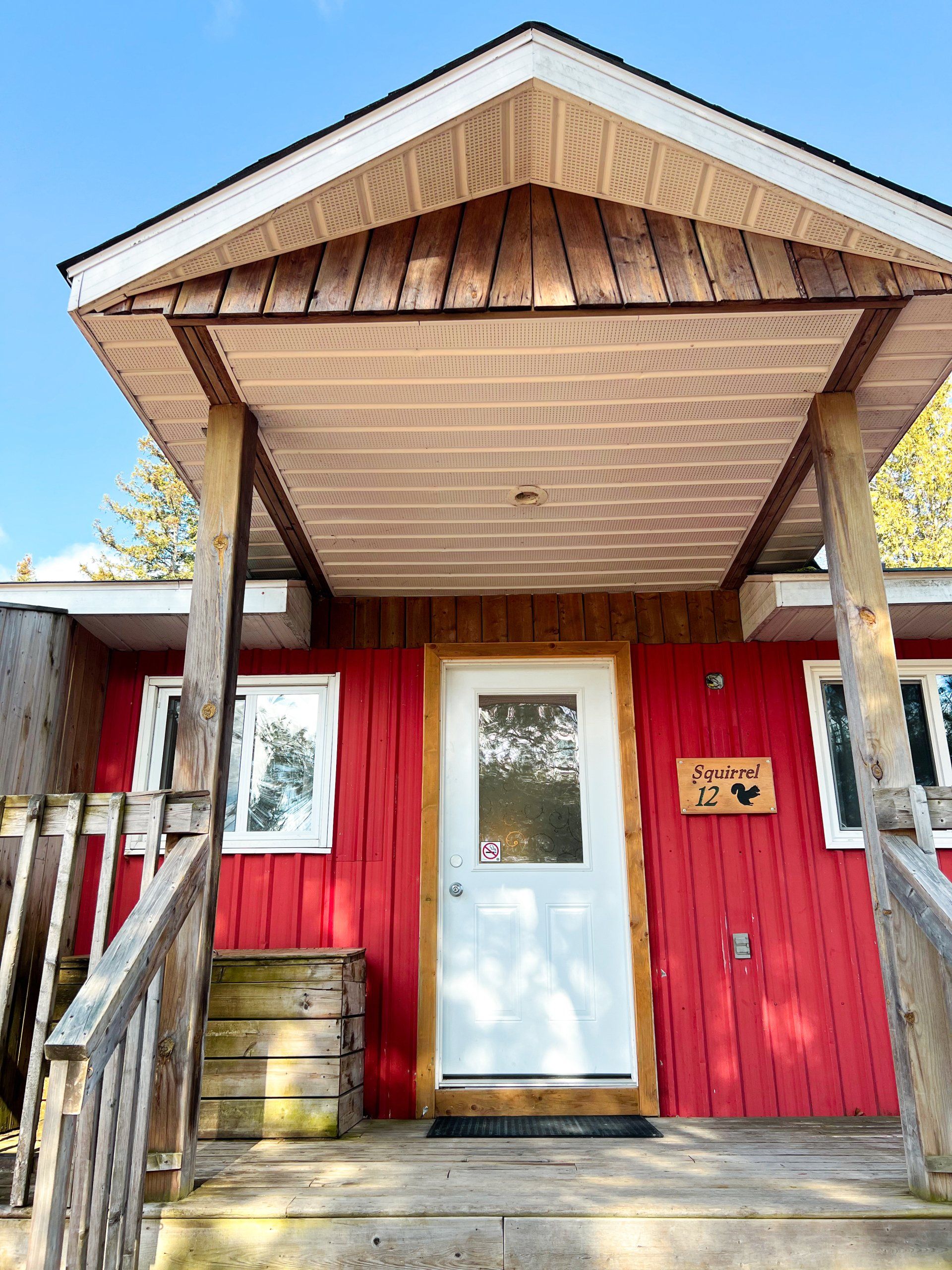 Red wooden cabin entrance with white door, small porch, and brown railing under a peaked roof.