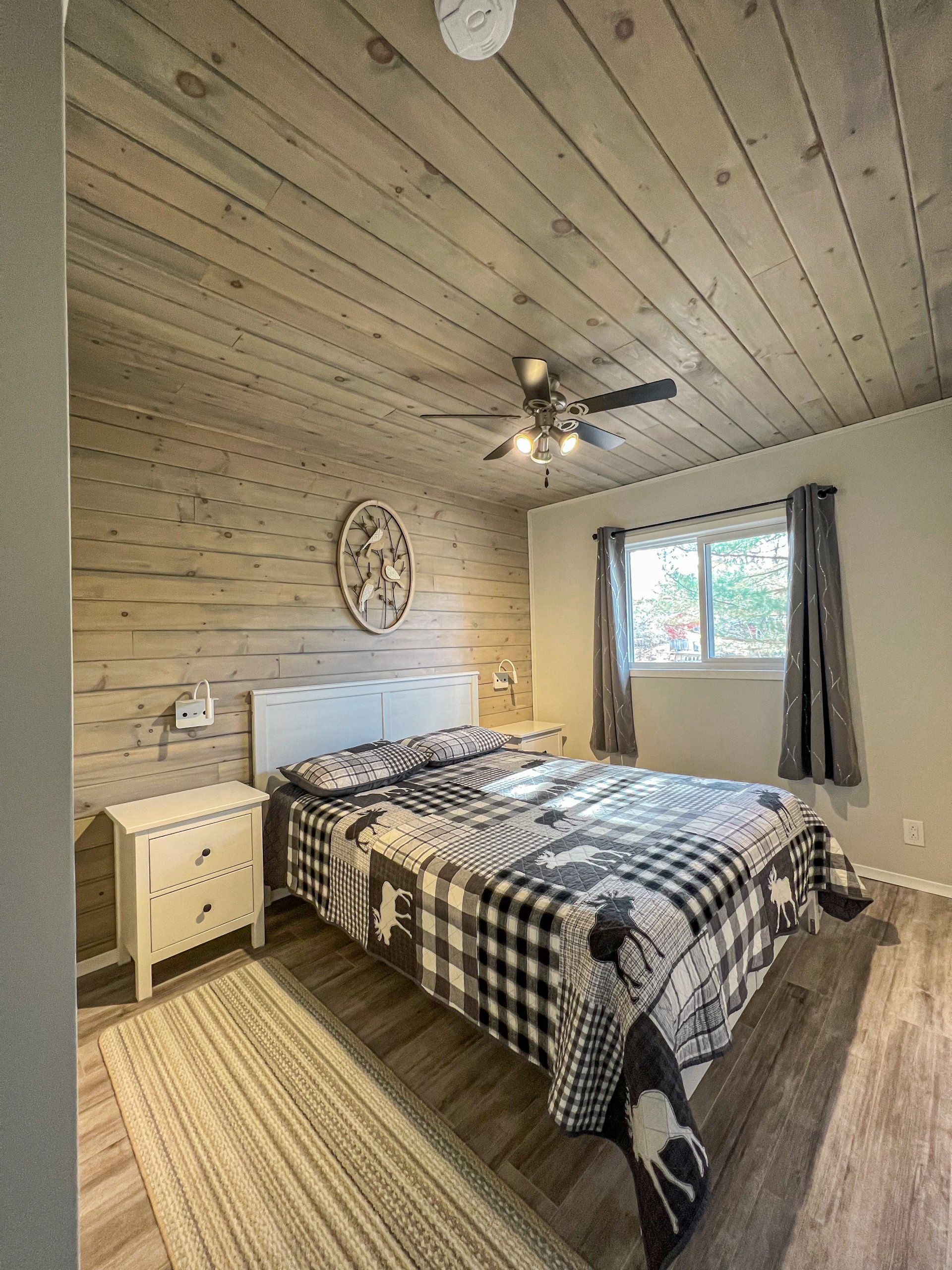 Cozy bedroom with plaid bedspread, white walls, wood ceiling, window, and ceiling fan.