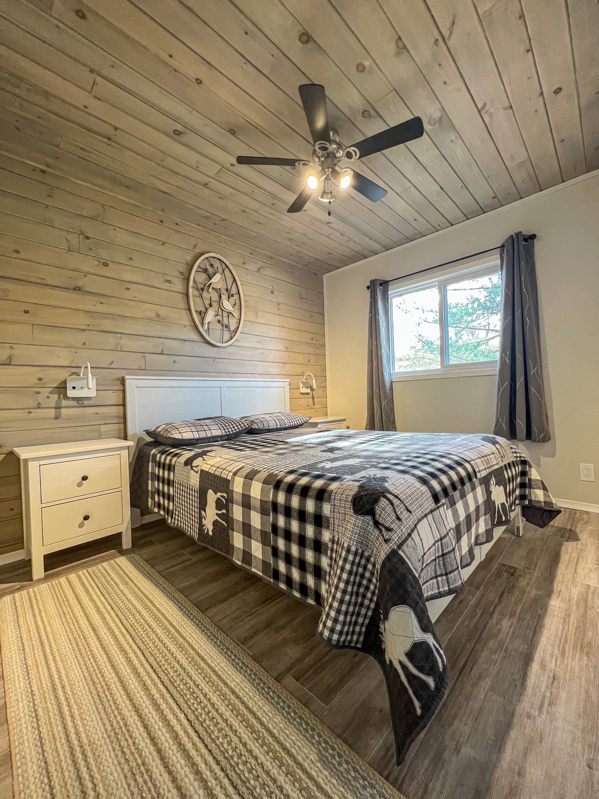 Cozy rustic bedroom with wood walls, plaid bedspread, nightstand, ceiling fan, and window with curtains