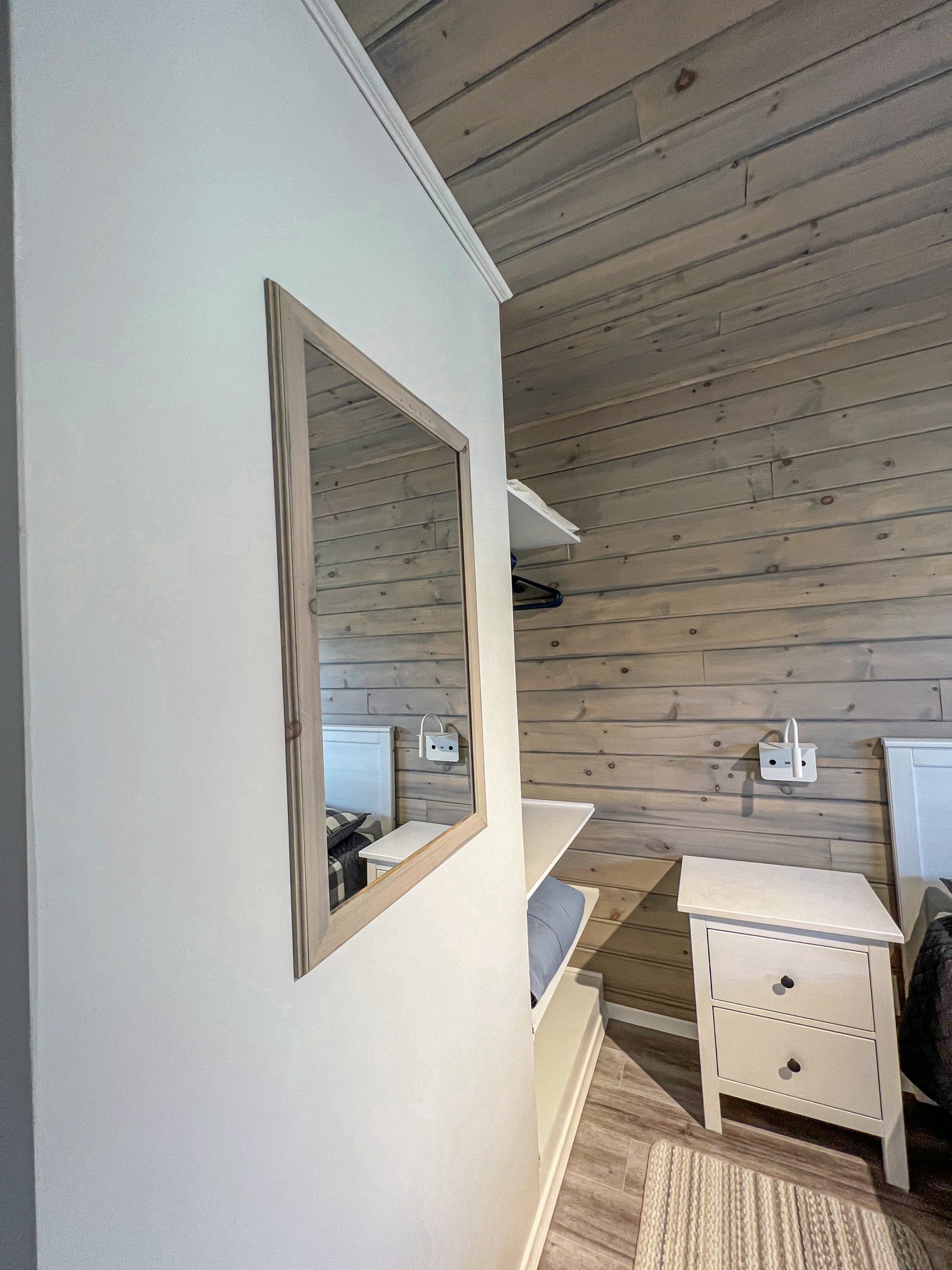 Rustic bedroom corner with a white wall mirror, wooden plank walls, and a small dresser by the bed.