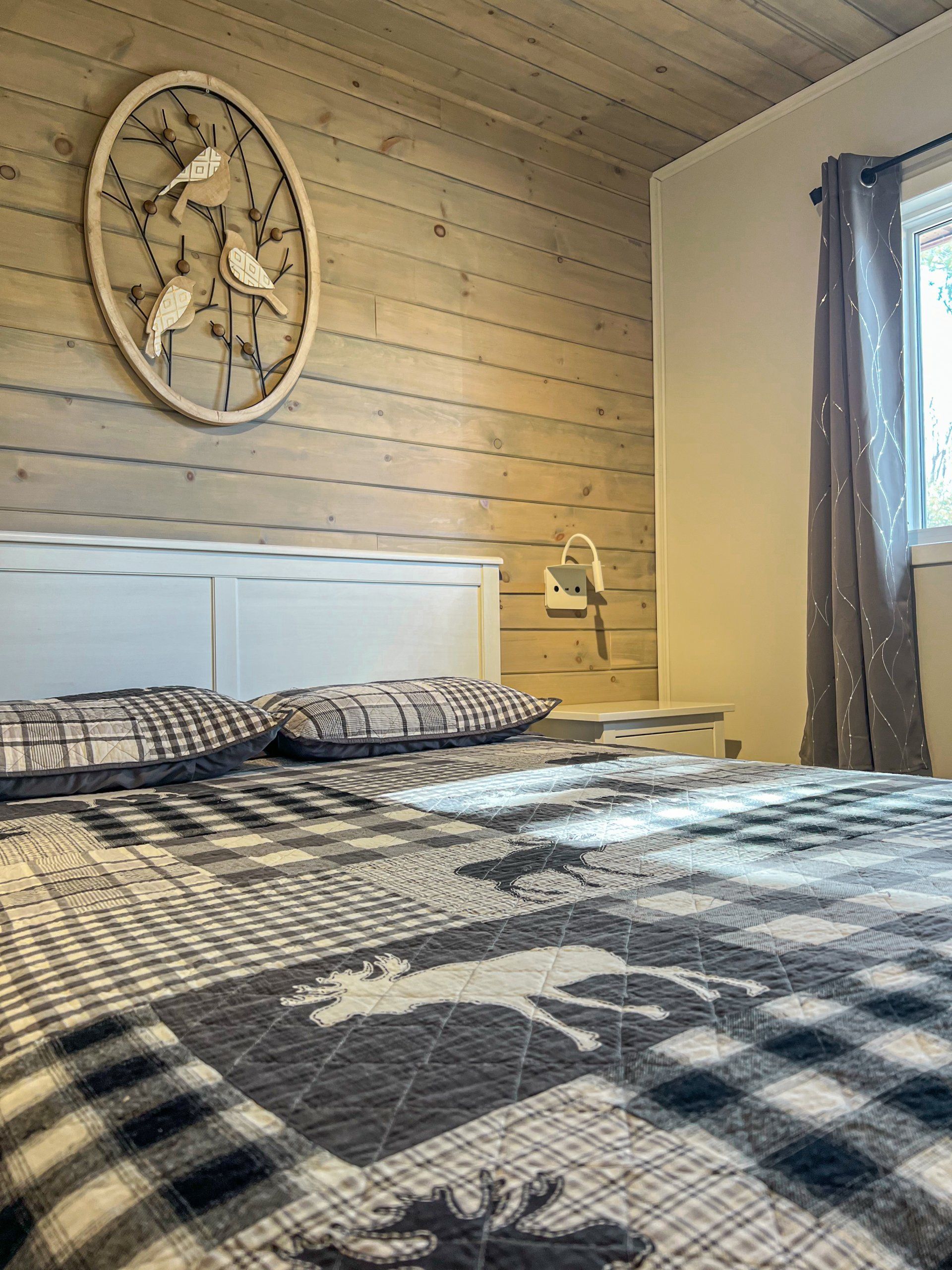 Cozy bedroom with plaid bedspread, white headboard, wood-paneled wall, and large rustic wall clock