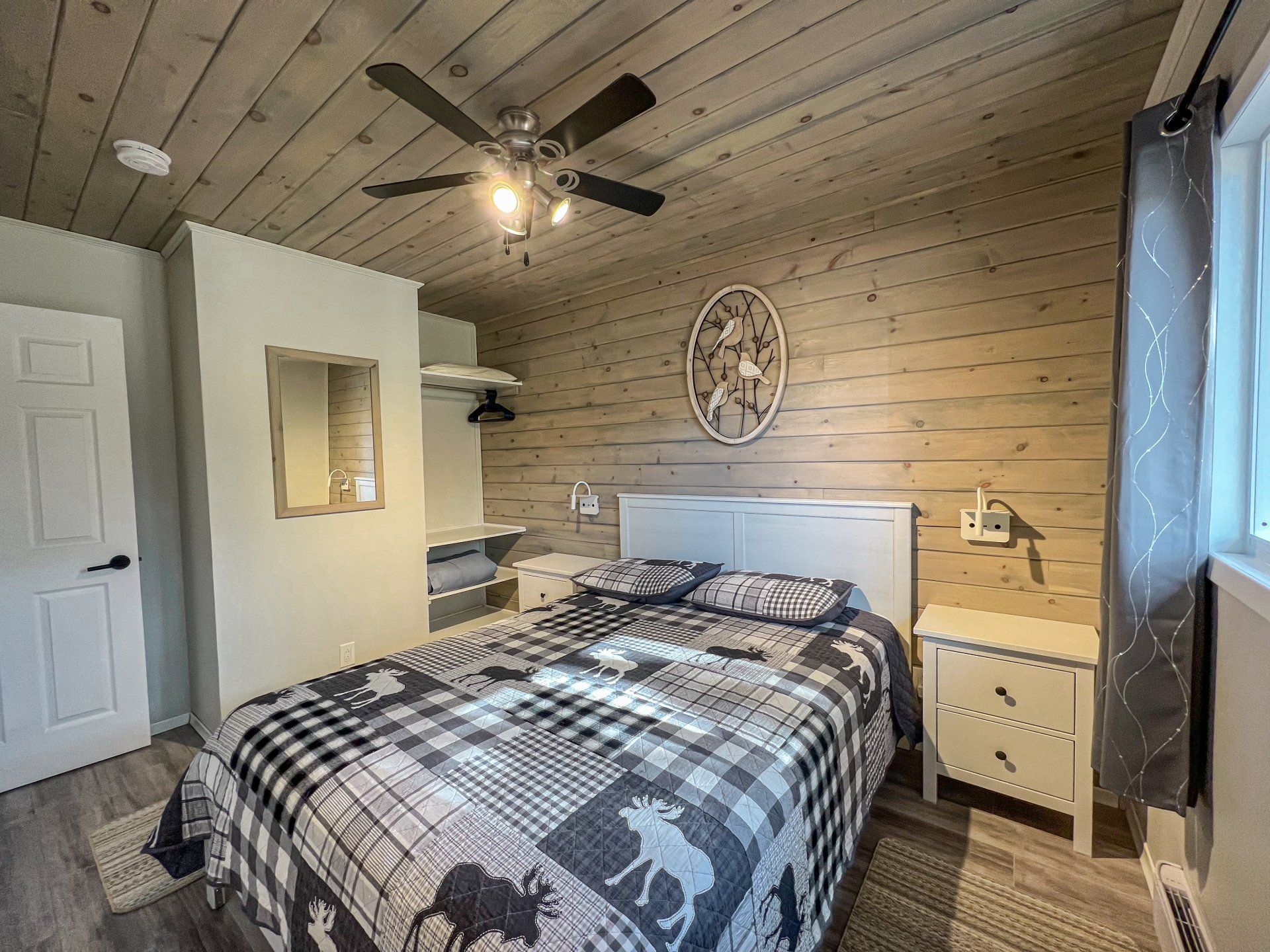 Cozy bedroom with a plaid bedspread, wood-paneled walls, ceiling fan, and window curtains