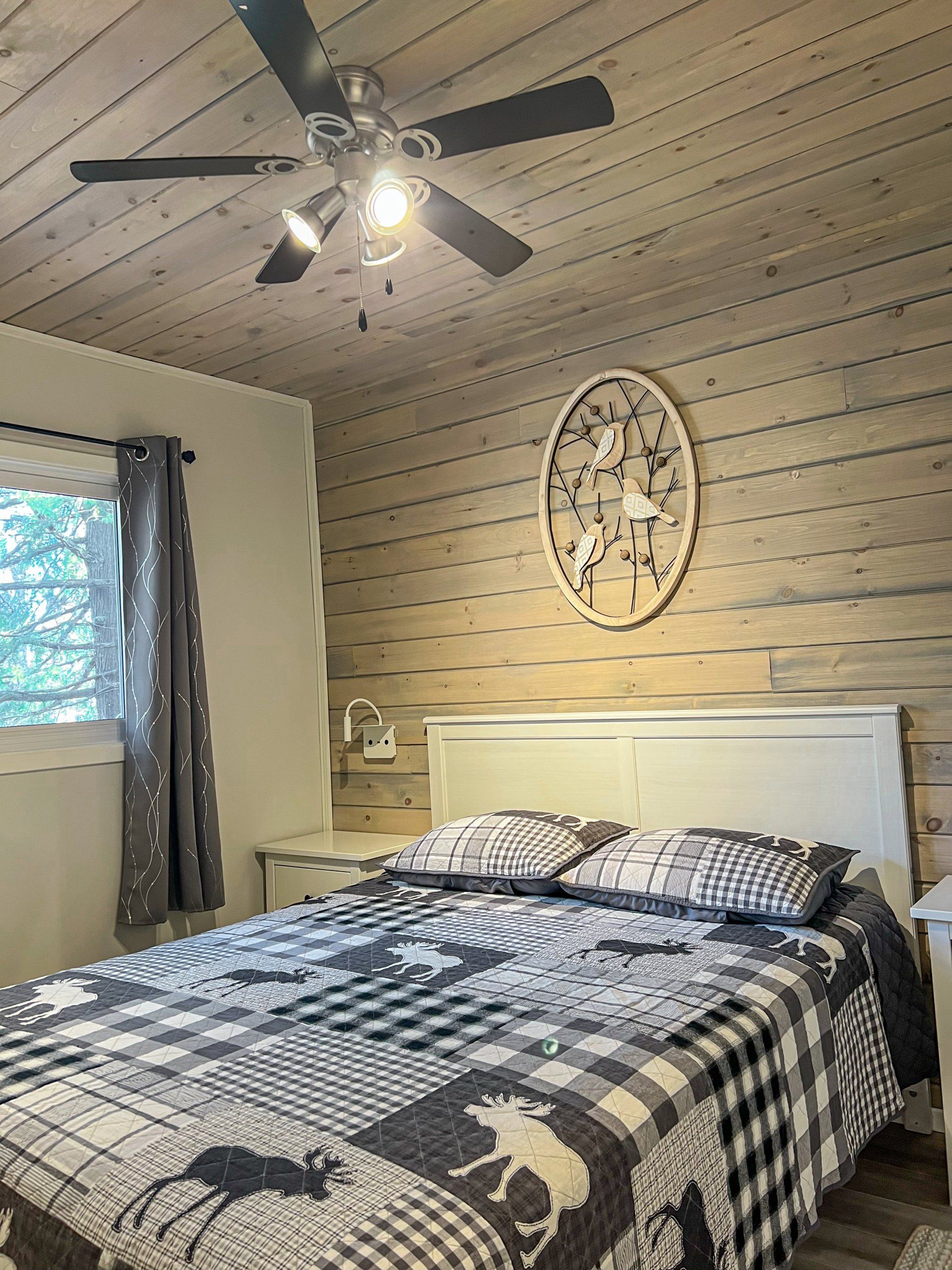 Bedroom with a bed, wood-paneled accent wall, ceiling fan light, window, and decorative wall art.