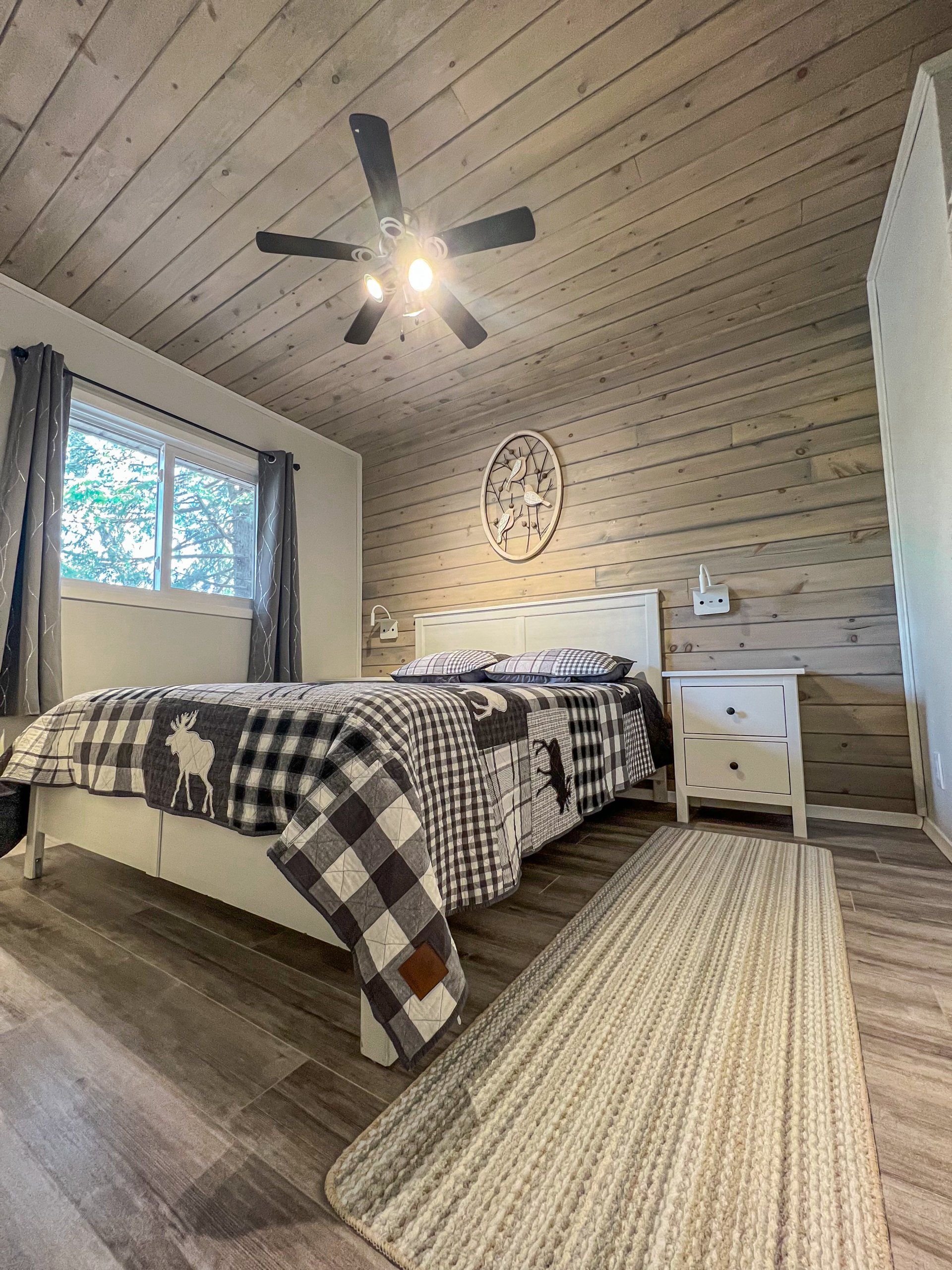 Cozy bedroom with plaid bedspread, woven rug, wood walls, ceiling fan, and window light