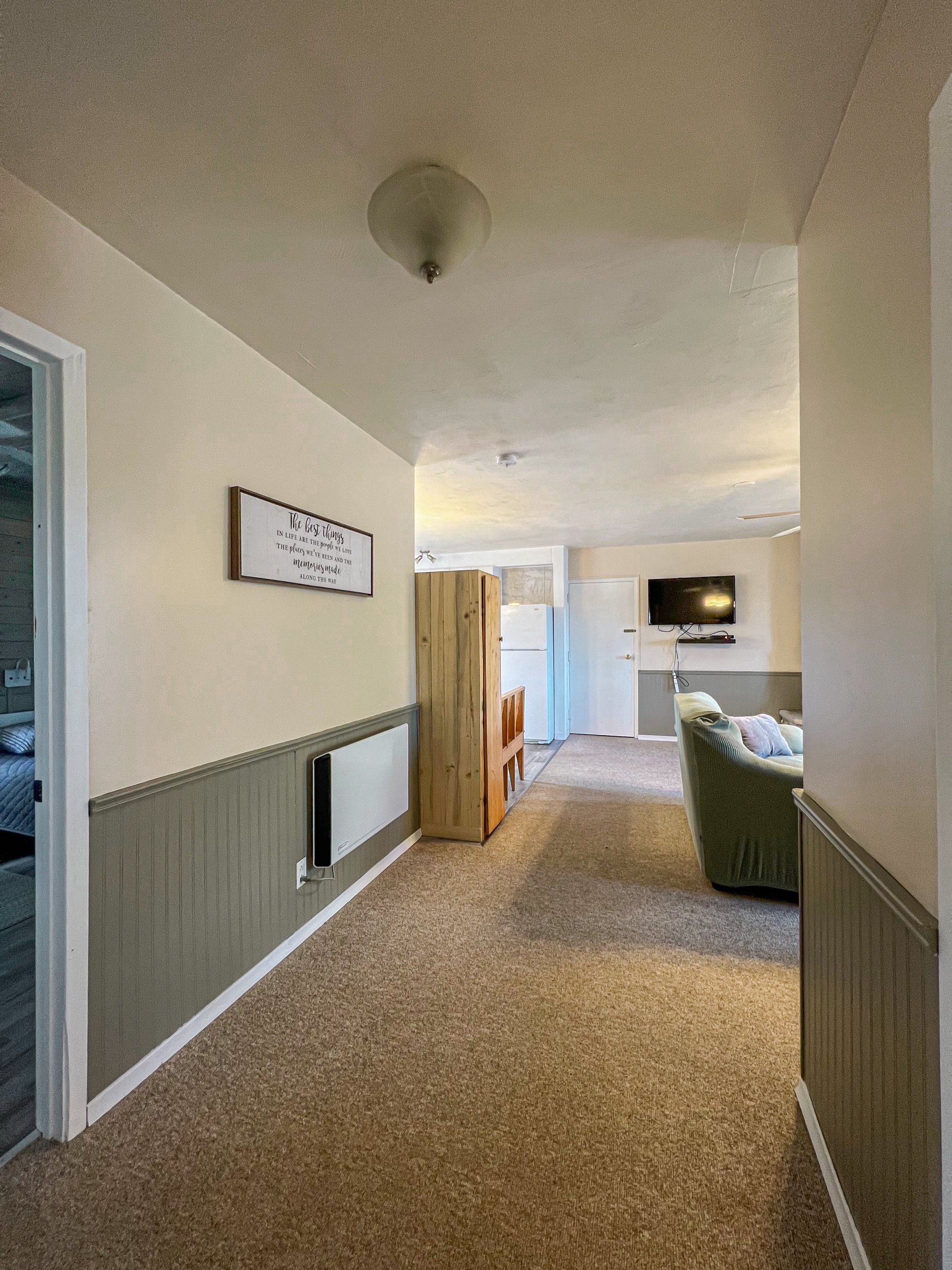 Narrow carpeted hallway with wainscoting, leading to a living room with a sofa and wall-mounted TV.