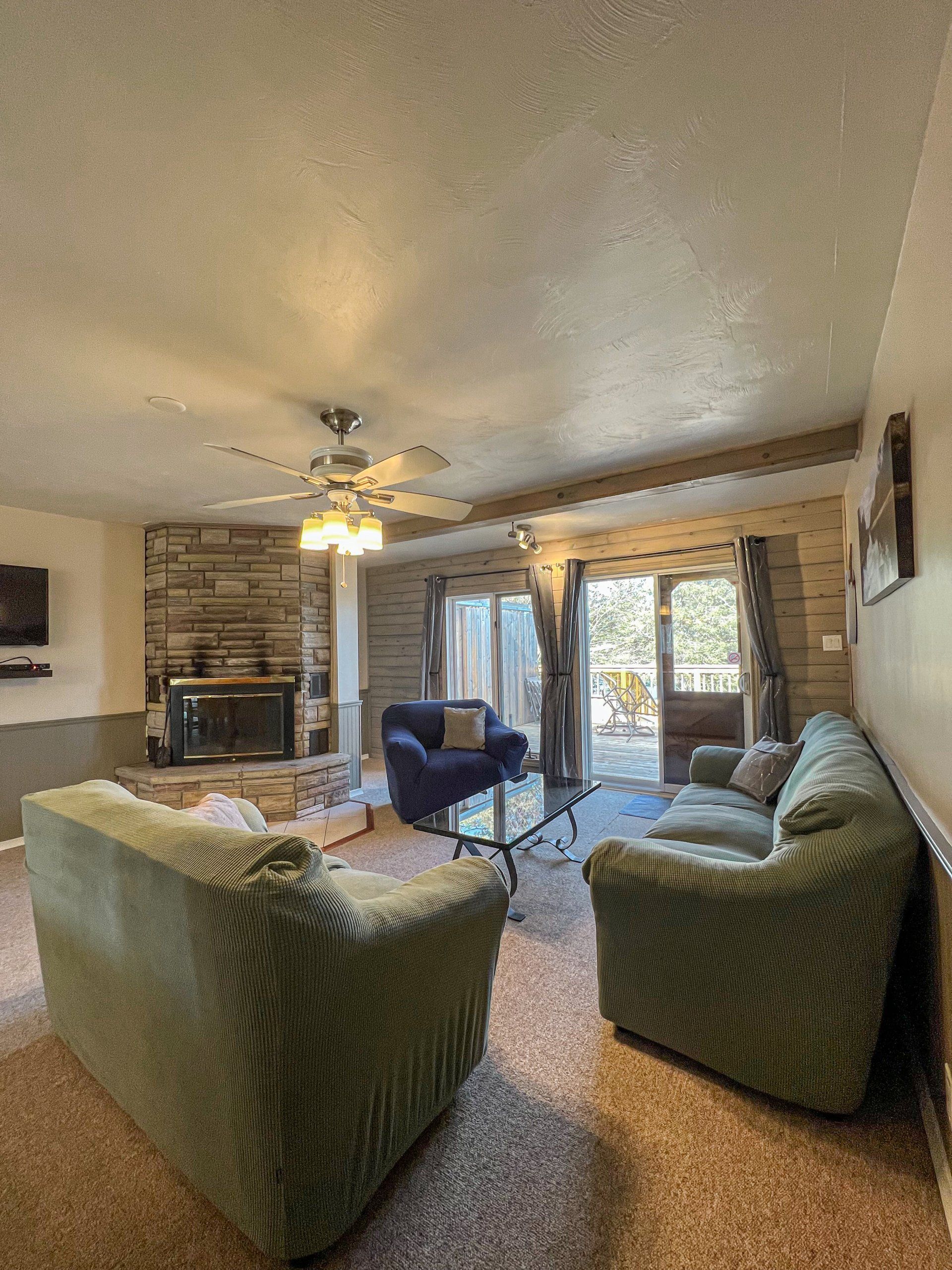 Cozy living room with green sofas, blue chair, stone fireplace, ceiling fan, and glass doors to a deck