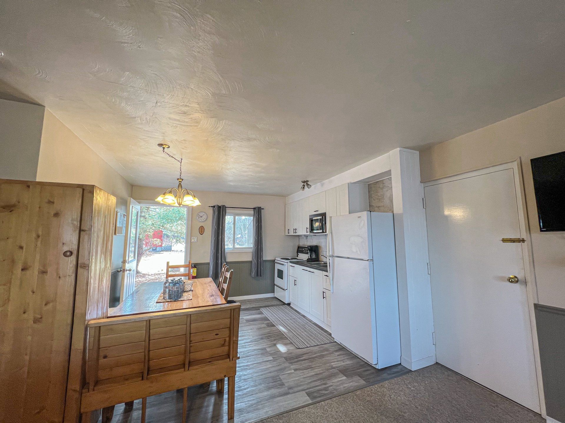 Small kitchen and dining area with white cabinets, wooden table, patterned runner, and a bright doorway at the back.