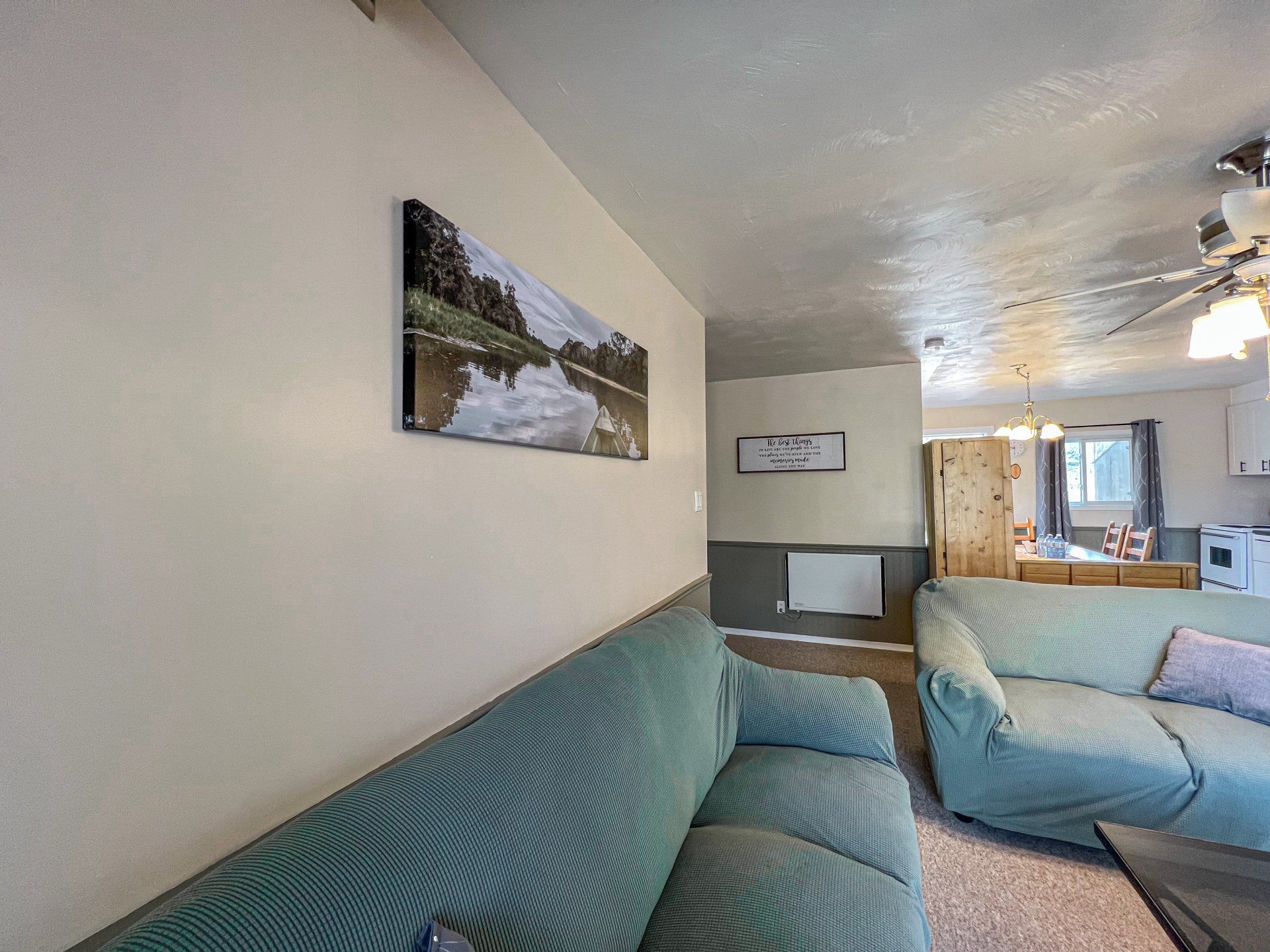 Small living room with teal couch, wall art, ceiling fan, and a doorway to a bright kitchen.