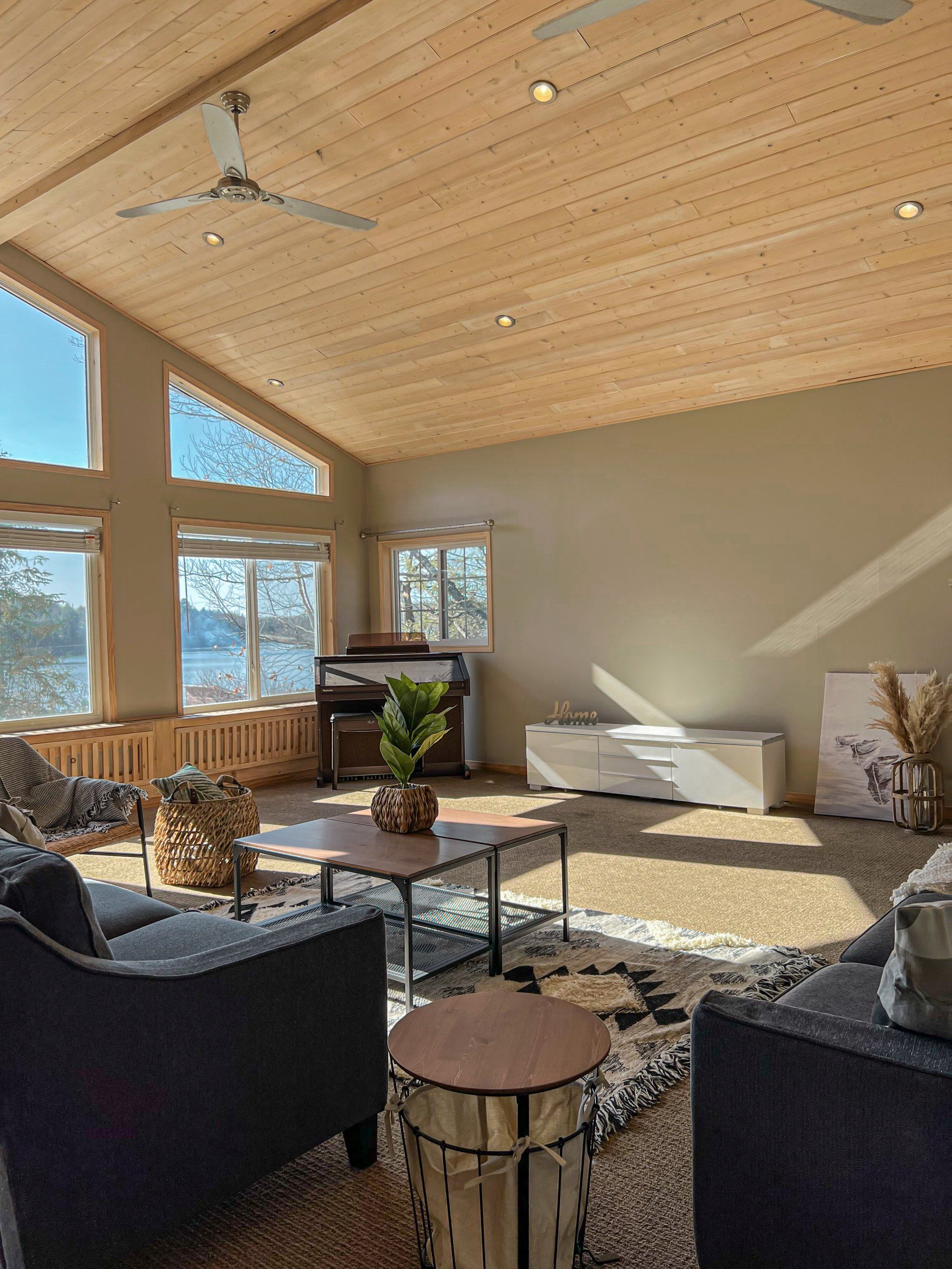 Sunlit living room with large windows, wood ceiling, dark chairs, coffee table, and a view outside