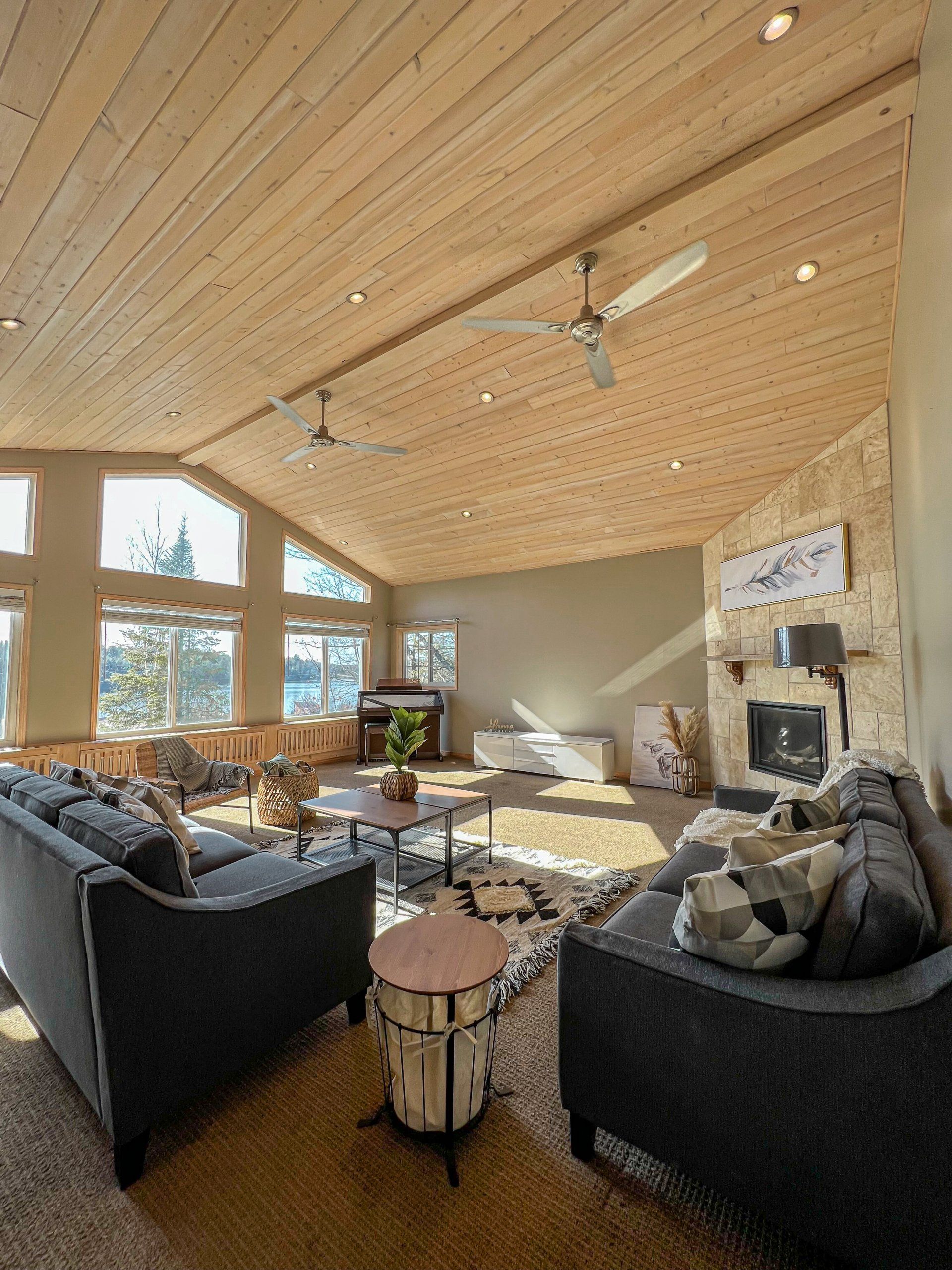 Bright living room with high wood ceiling, large windows, gray sofas, and stone fireplace.