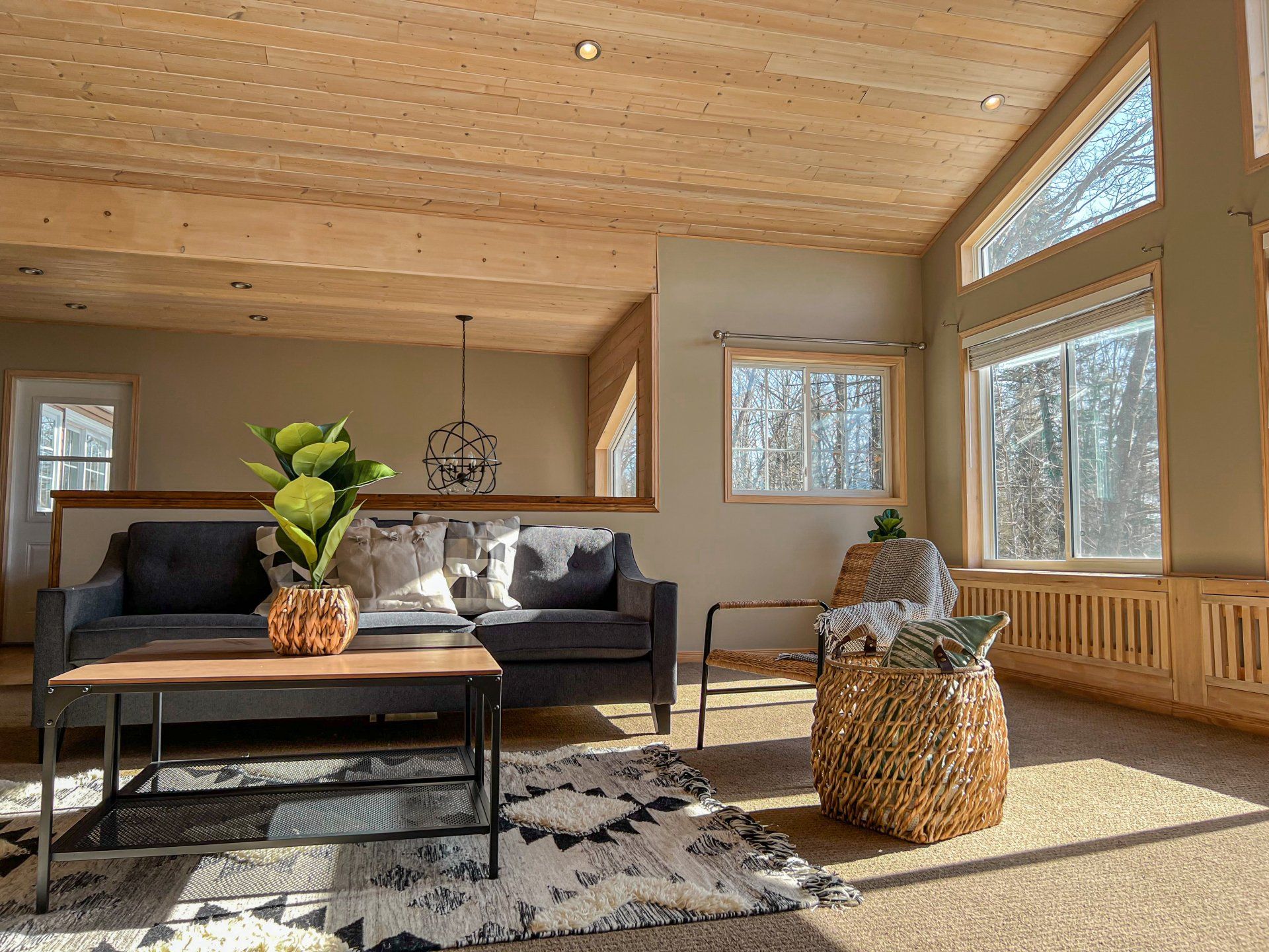 Bright sunlit living room with gray sofa, coffee table, woven ottomans, and large windows.
