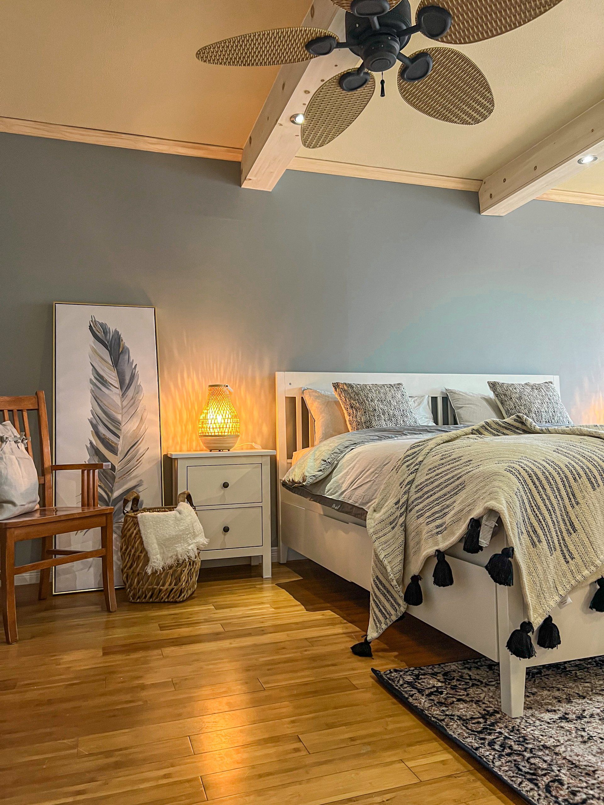 Cozy bedroom with bed, white dresser, warm lamp, wood floor, and ceiling fan on a gray wall