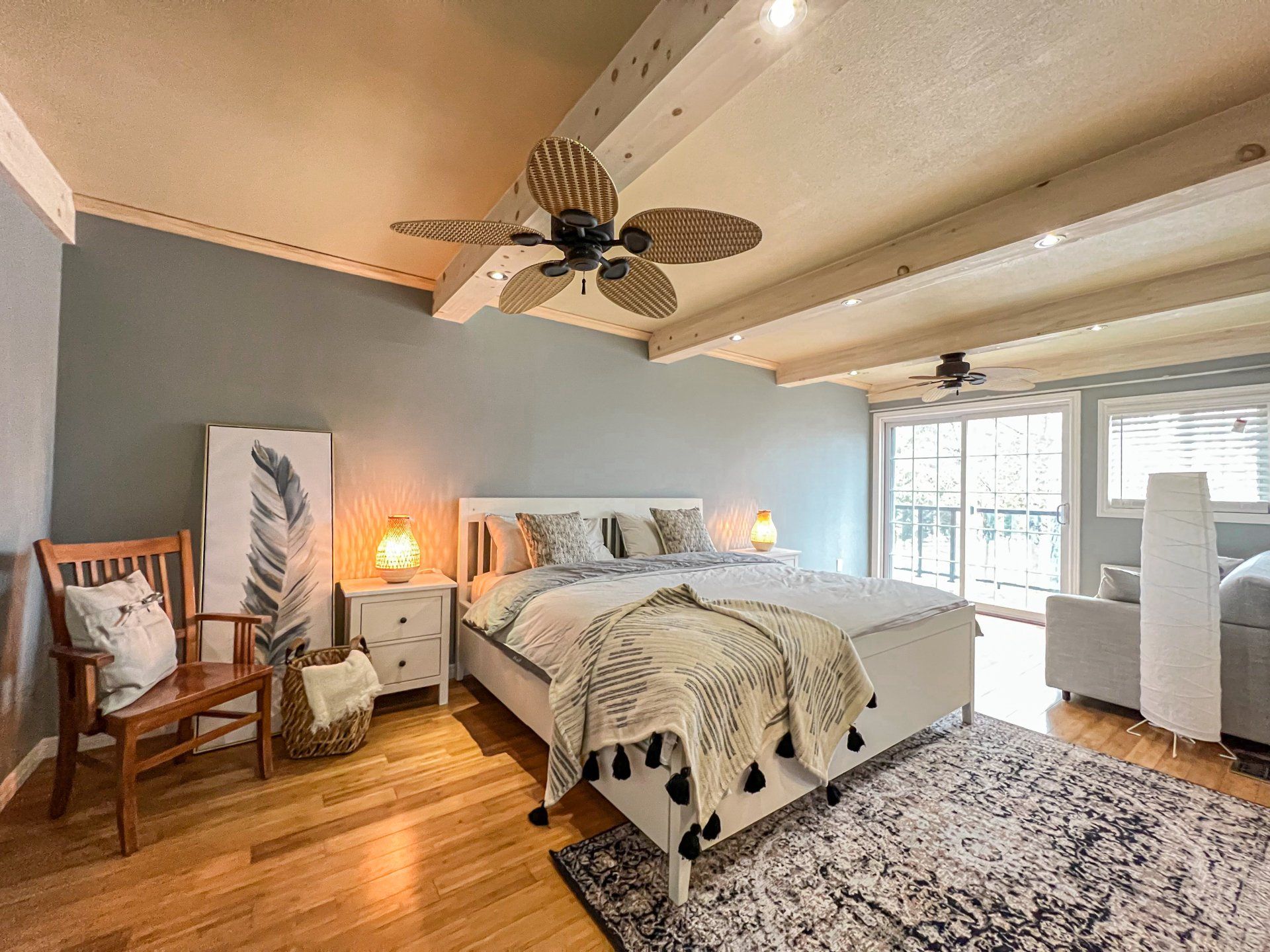 Cozy bedroom with a bed, wooden chair, gray accent wall, rug, and ceiling fan near bright windows