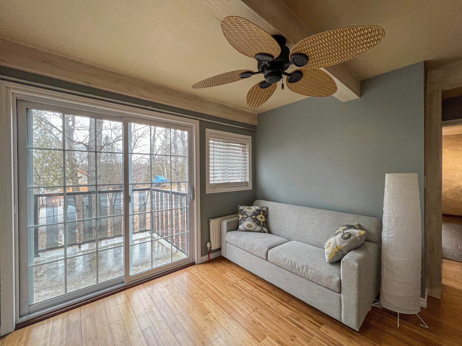 Bright sunroom with sliding glass doors, light wood floor, gray sofa, and ceiling fan with leaf blades