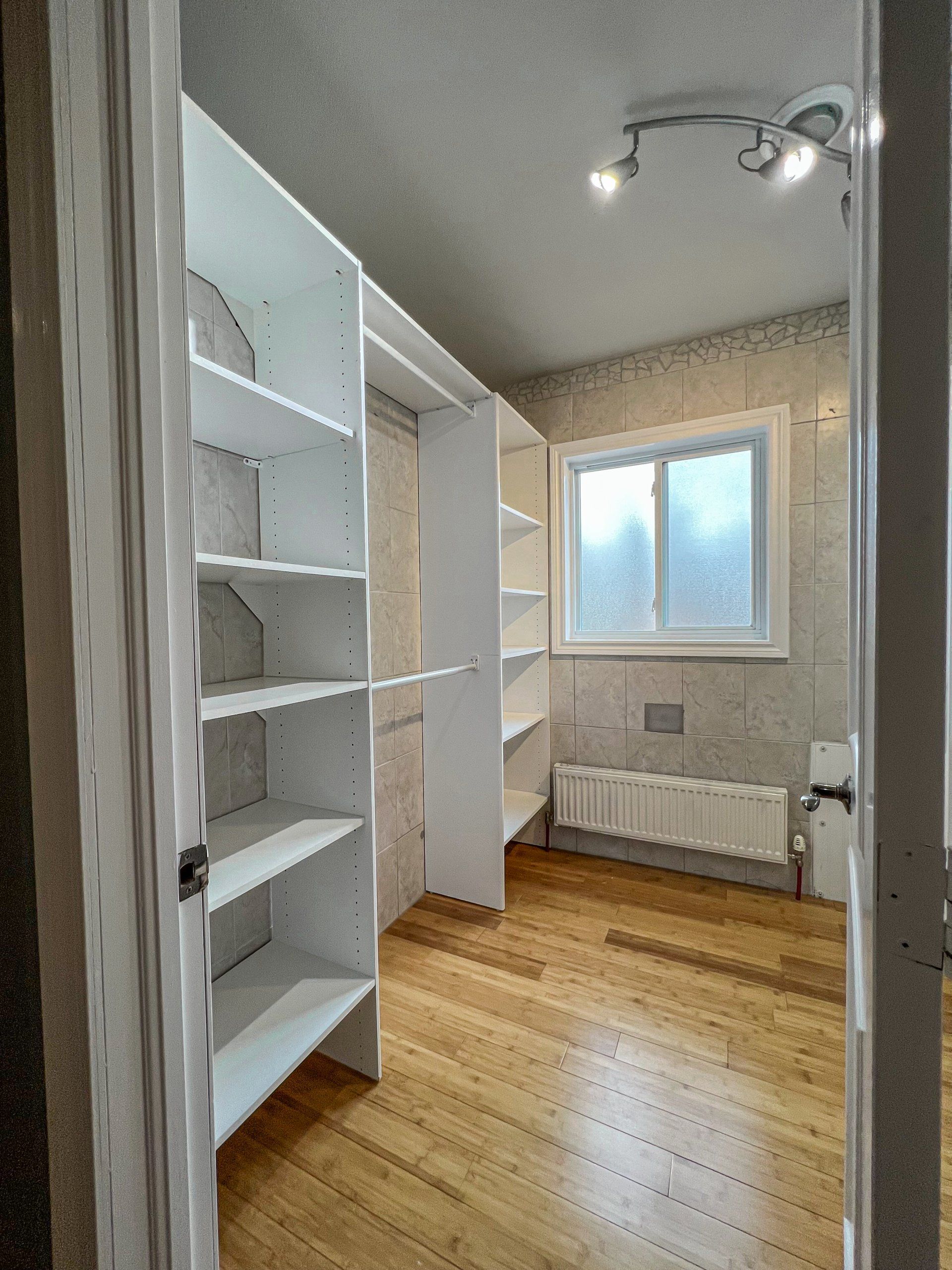 Empty walk-in closet with white shelves, wood floor, window, radiator, and ceiling lights.