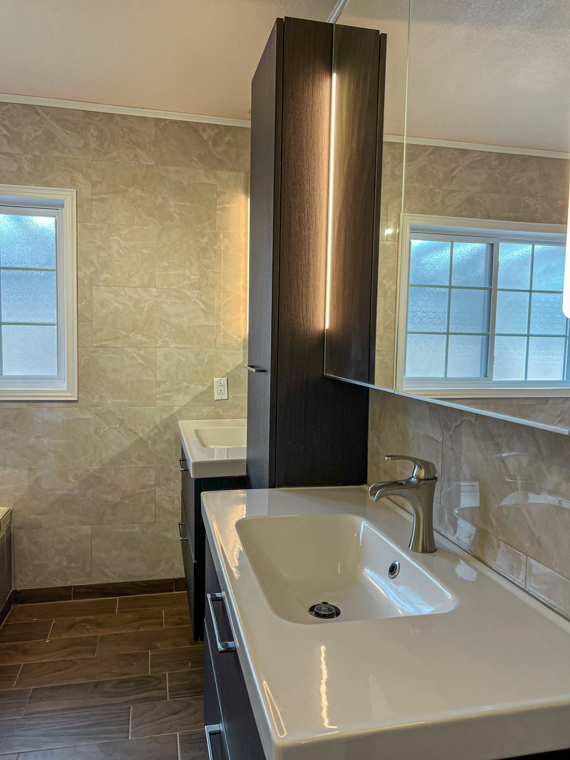 Modern kitchen with white sink, stone countertop, tall dark cabinet, and windows overlooking a tiled wall