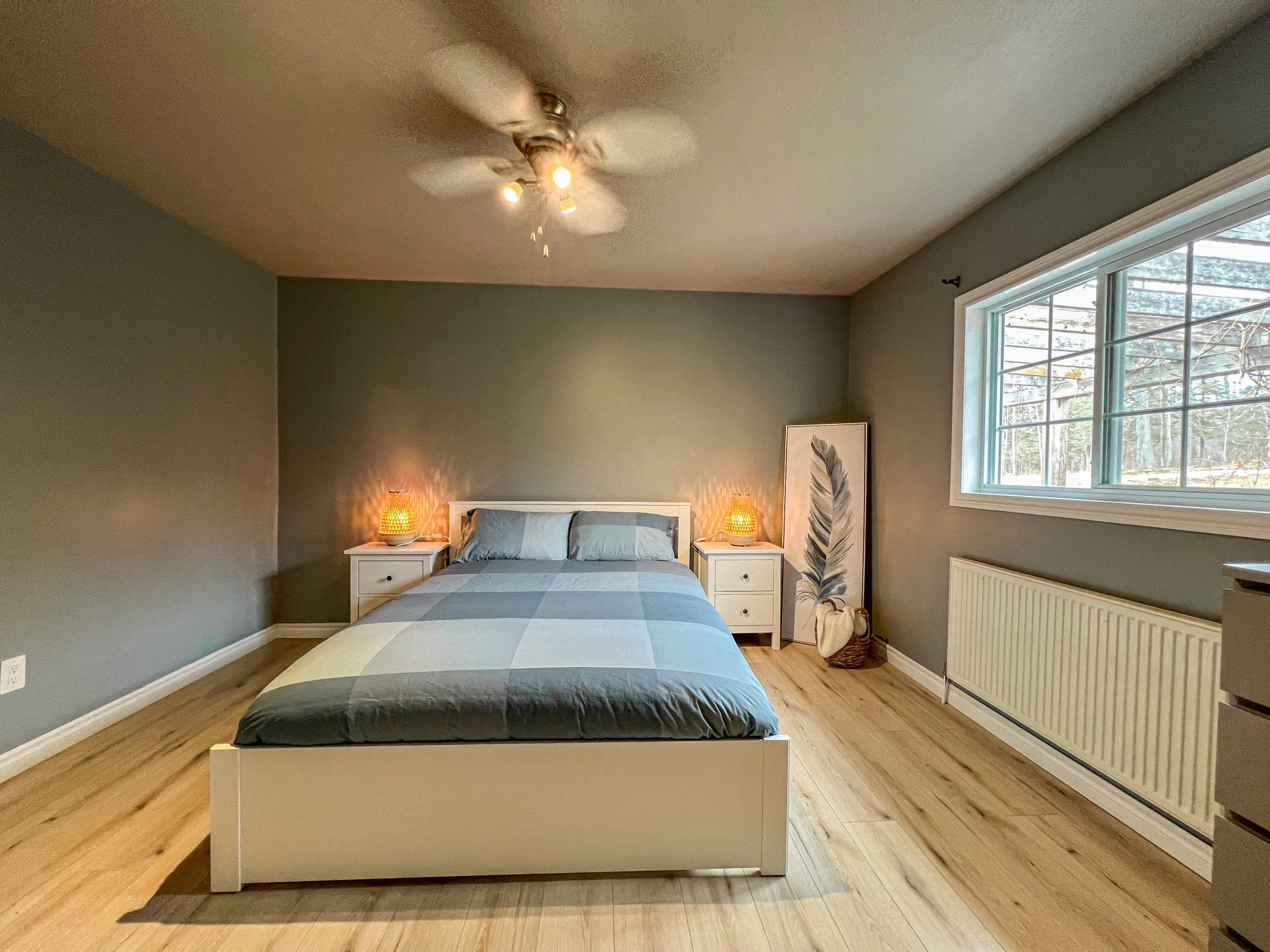 Bedroom with gray bed, warm lamp, wood floor, window, and ceiling fan in a minimalist setting