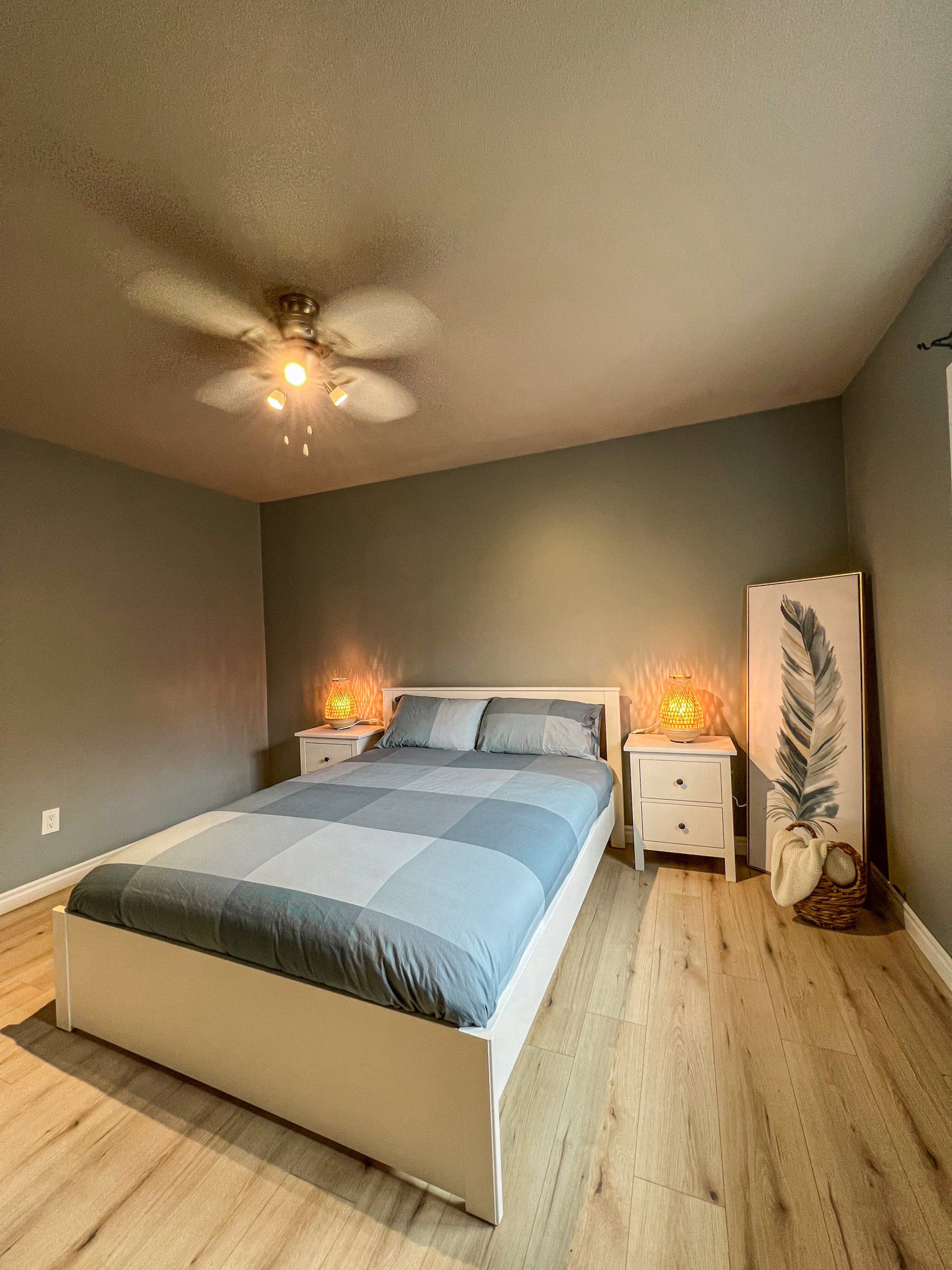 Modern bedroom with gray walls, a bed, nightstands, warm lamps, and a ceiling fan
