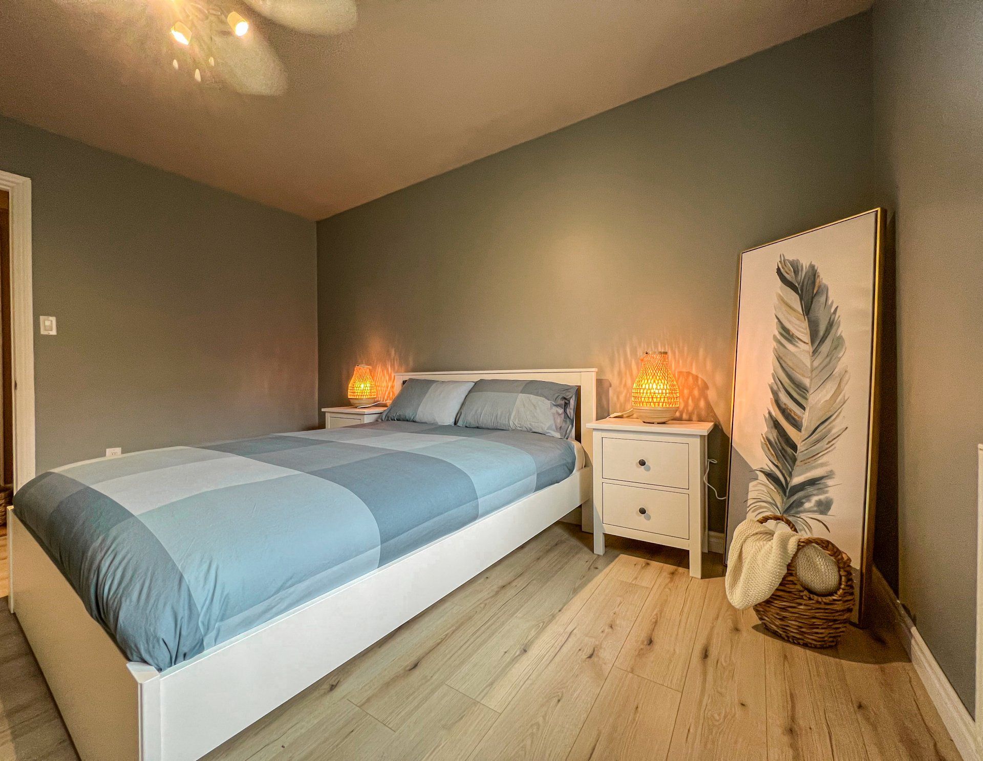 Modern bedroom with blue bedspread, gray walls, wooden floor, nightstands, lamp, and feather wall art.