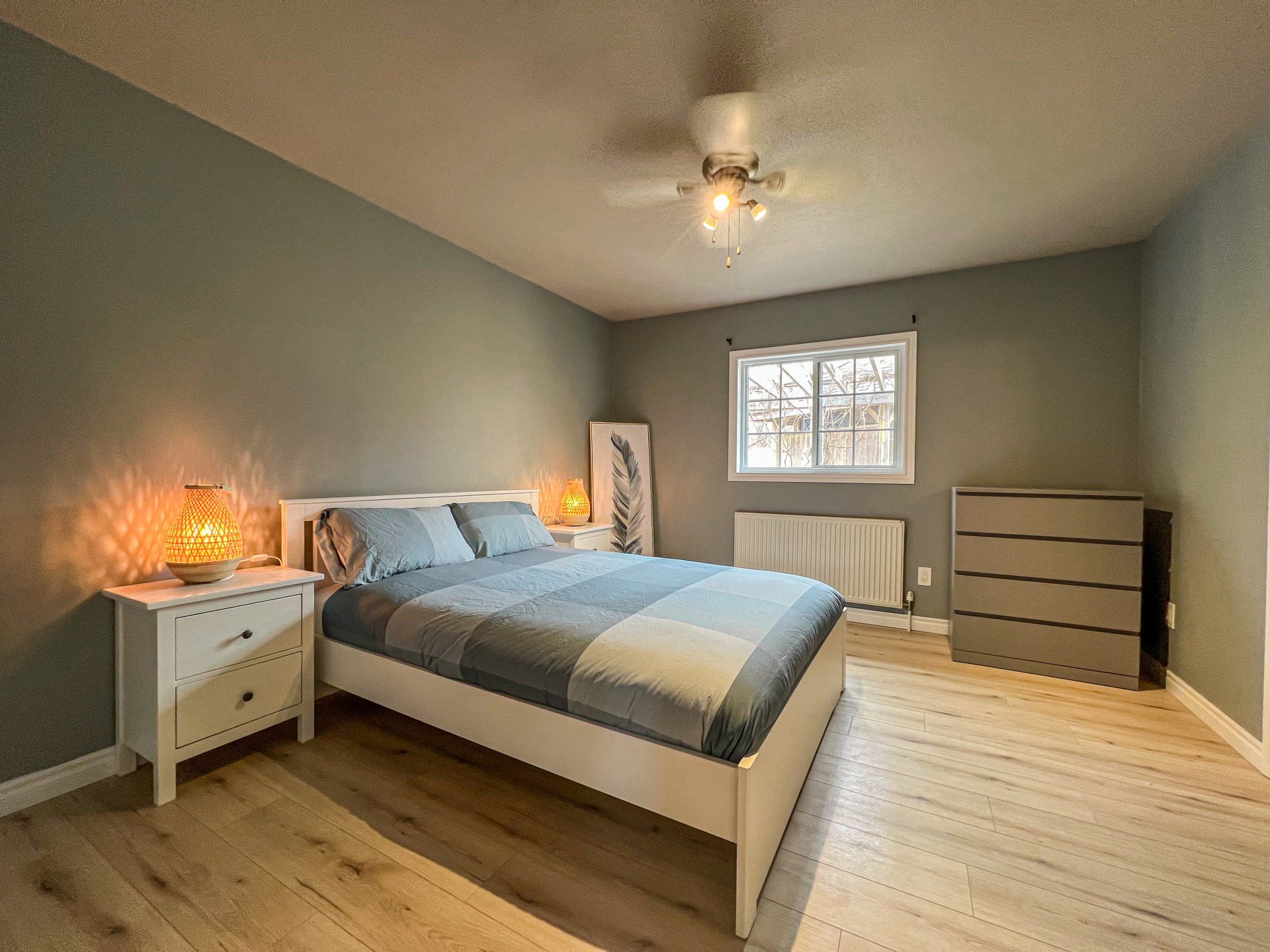 Bright bedroom with blue bedding, white dresser and nightstand, wood floor, and a window with blinds.