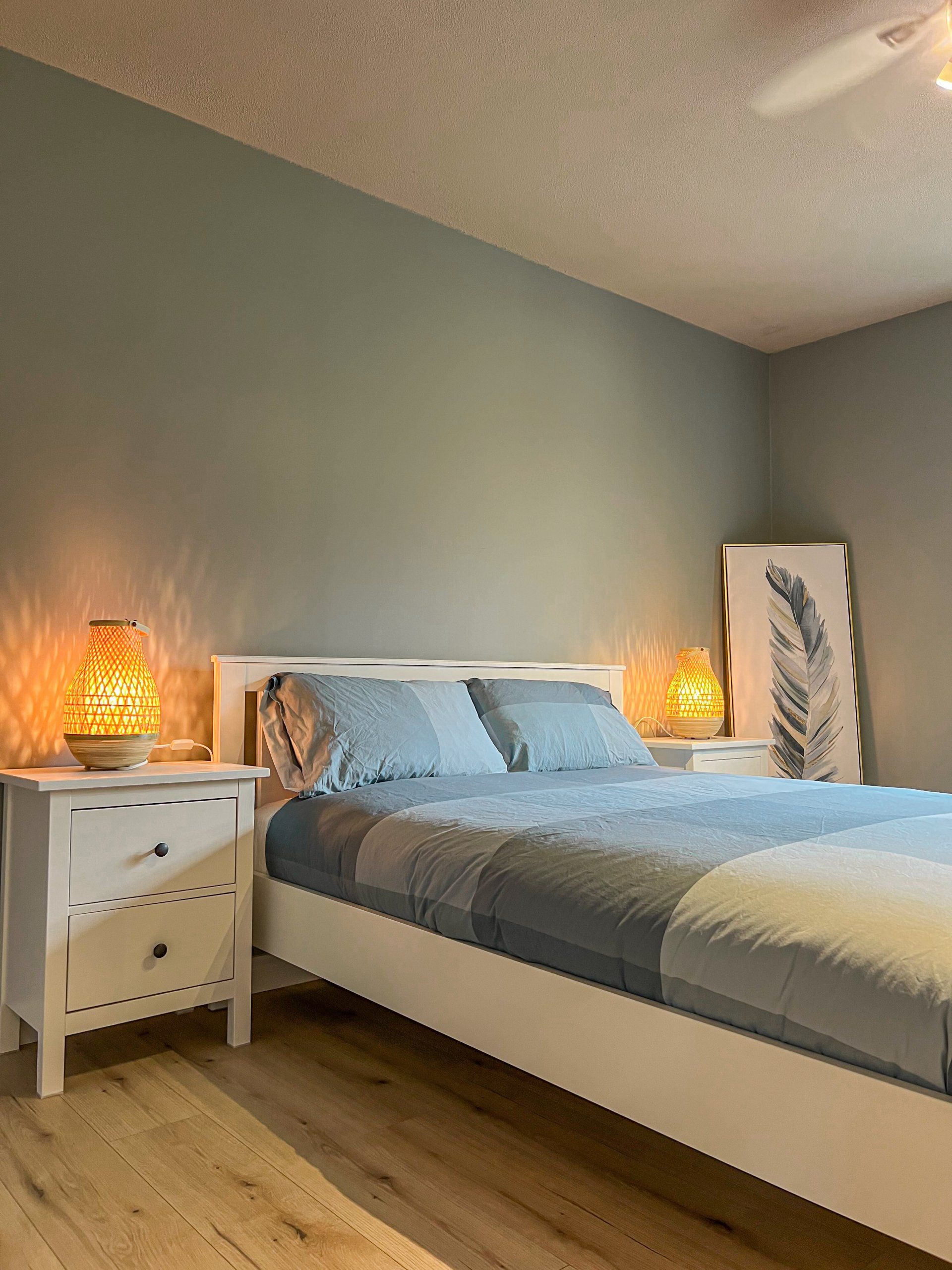 Cozy bedroom with a neatly made bed, soft lamp lighting, gray walls, and wood floors