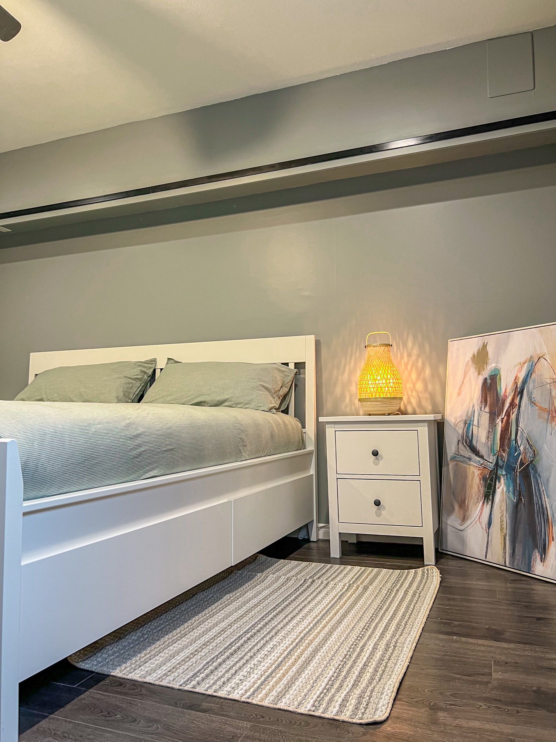 Modern bedroom with white bed, gray walls, nightstand lamp, rug, and leaning abstract art.