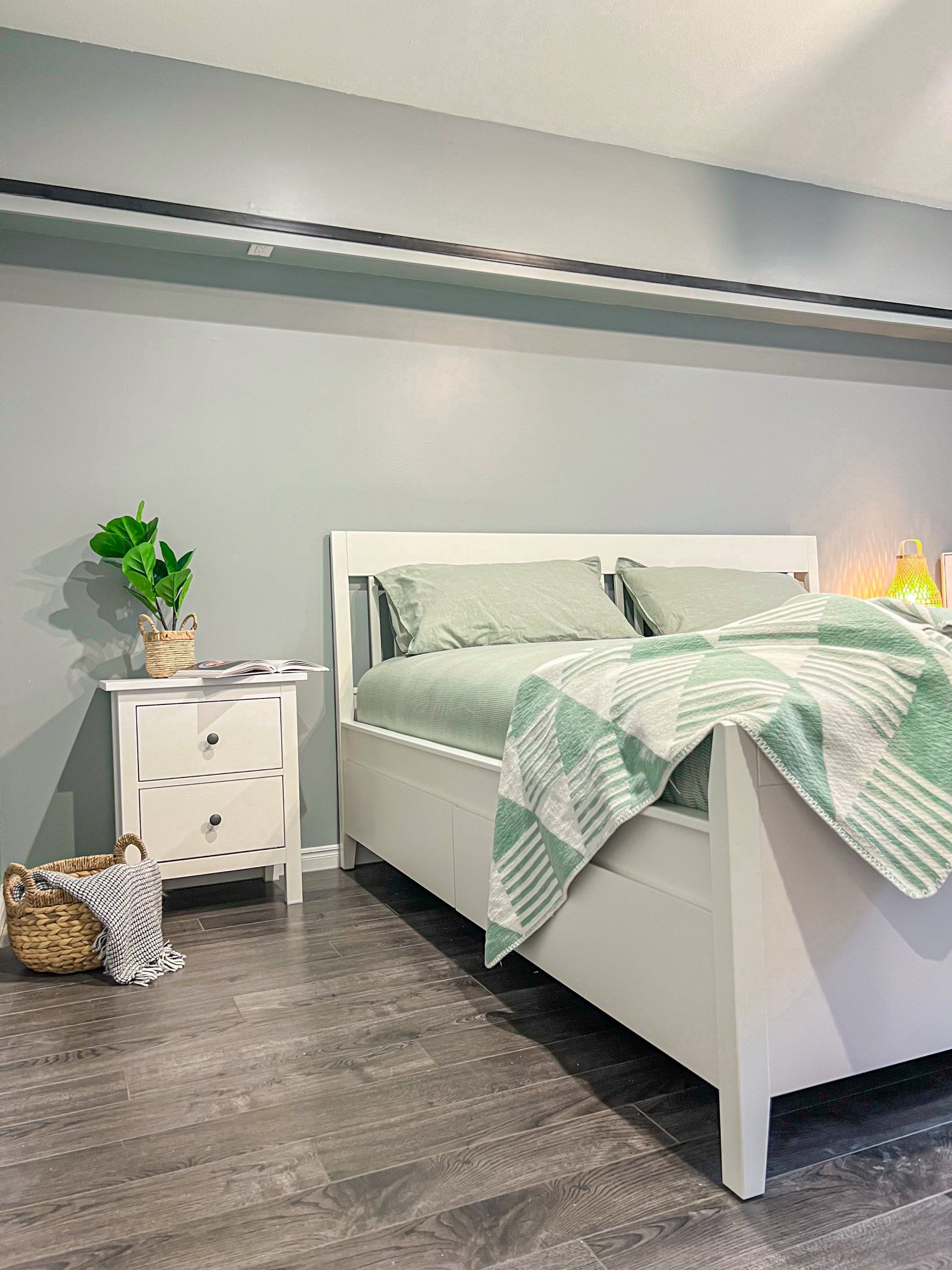 Bright bedroom with a white bed, mint bedding, nightstand, plant, and gray wood floor.