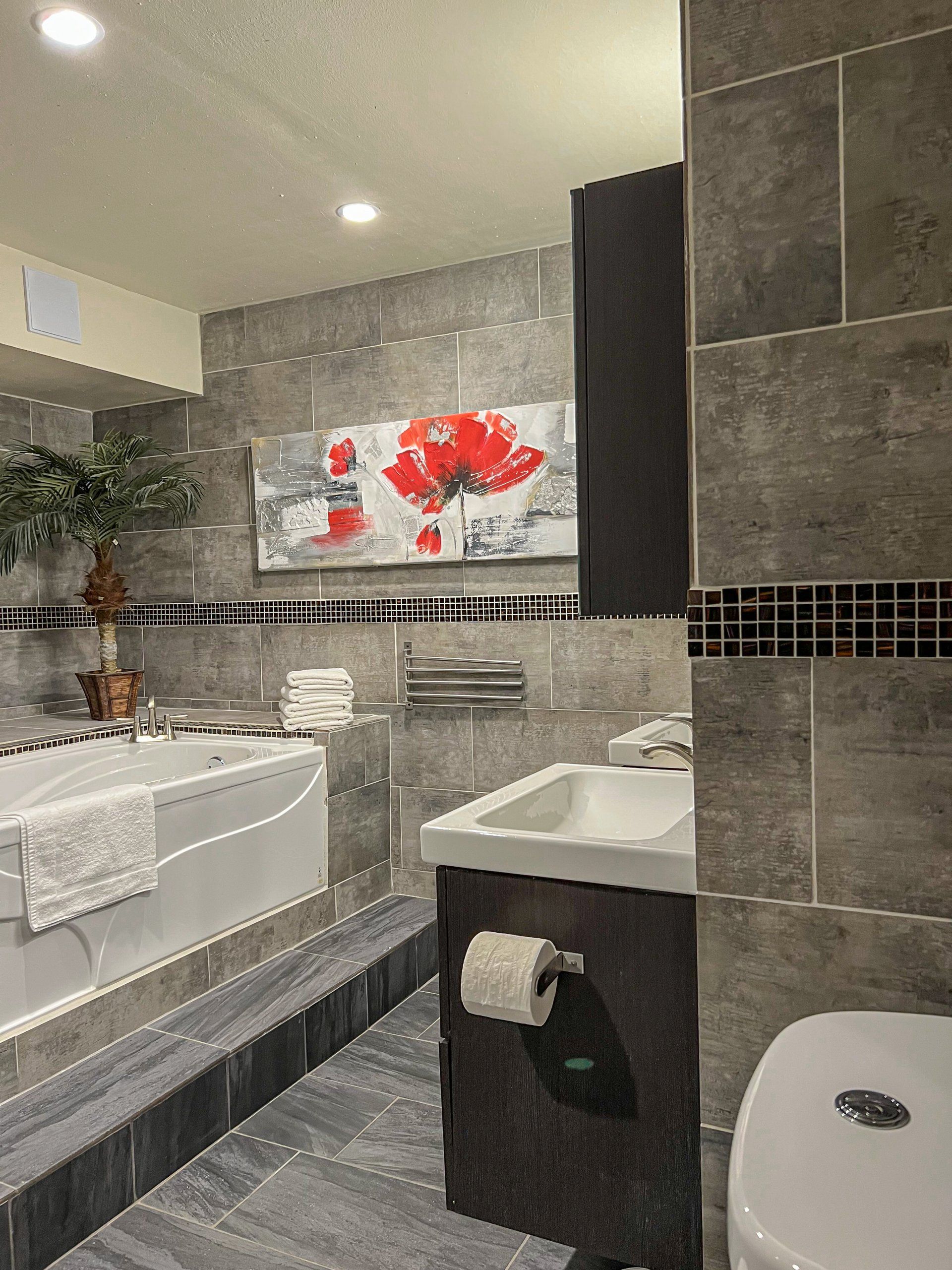 Modern gray-tiled bathroom with a white sink, bathtub, toilet, and red flower wall art