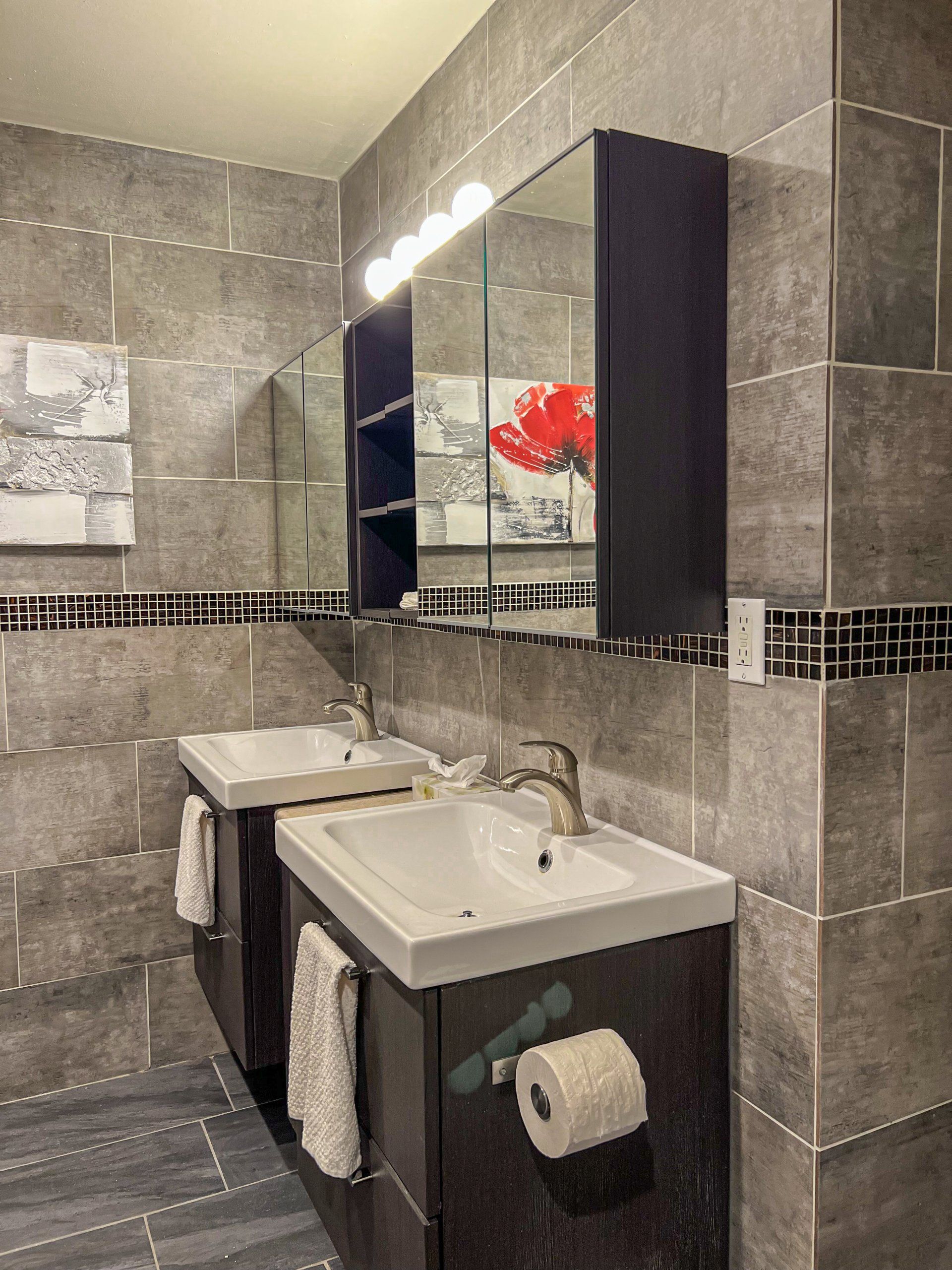 Modern tiled bathroom with sink, mirror cabinet, towel, toilet paper holder, and wall art.